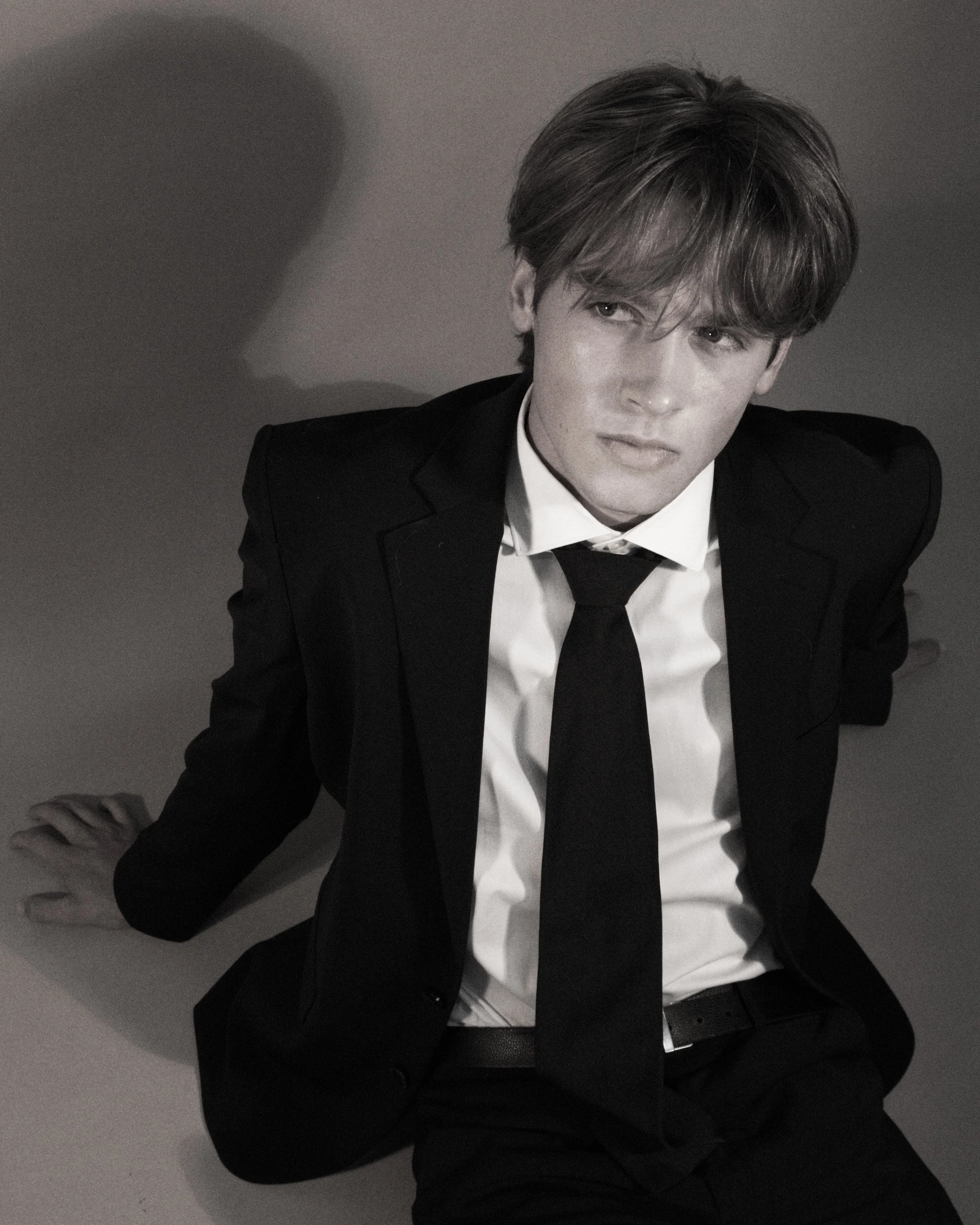 Black and white photo of a young man with styled hair, wearing a suit, sitting on a floor with a plain background, looking slightly to the side.