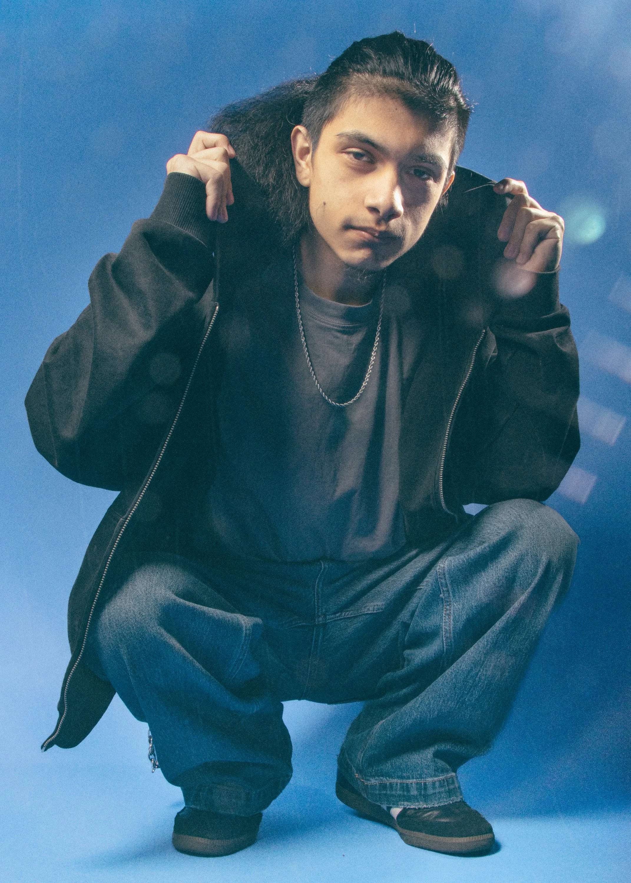 A person wearing a black jacket, gray t-shirt, blue jeans, and black shoes, crouching with a blue background.