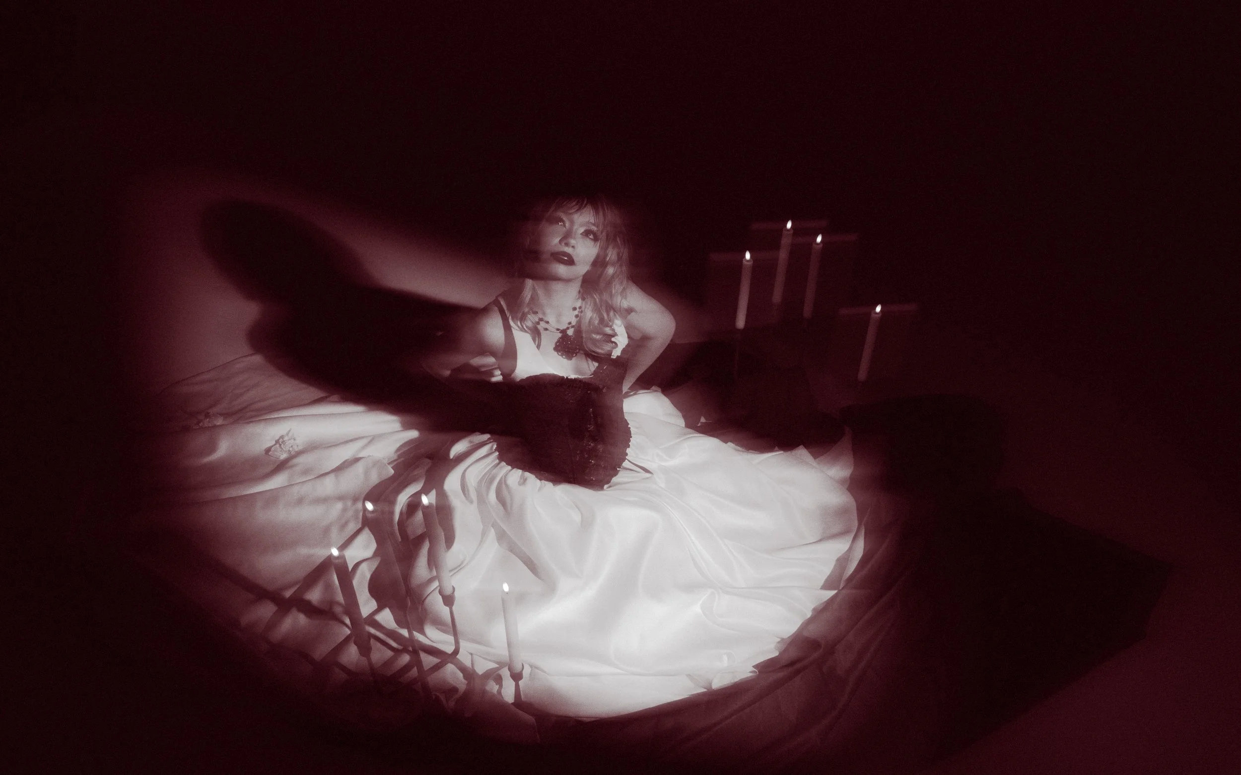 A woman with wavy hair and dark lipstick sitting on a bed in a dimly lit room, surrounded by lit candles and fairy lights, with a dark background.