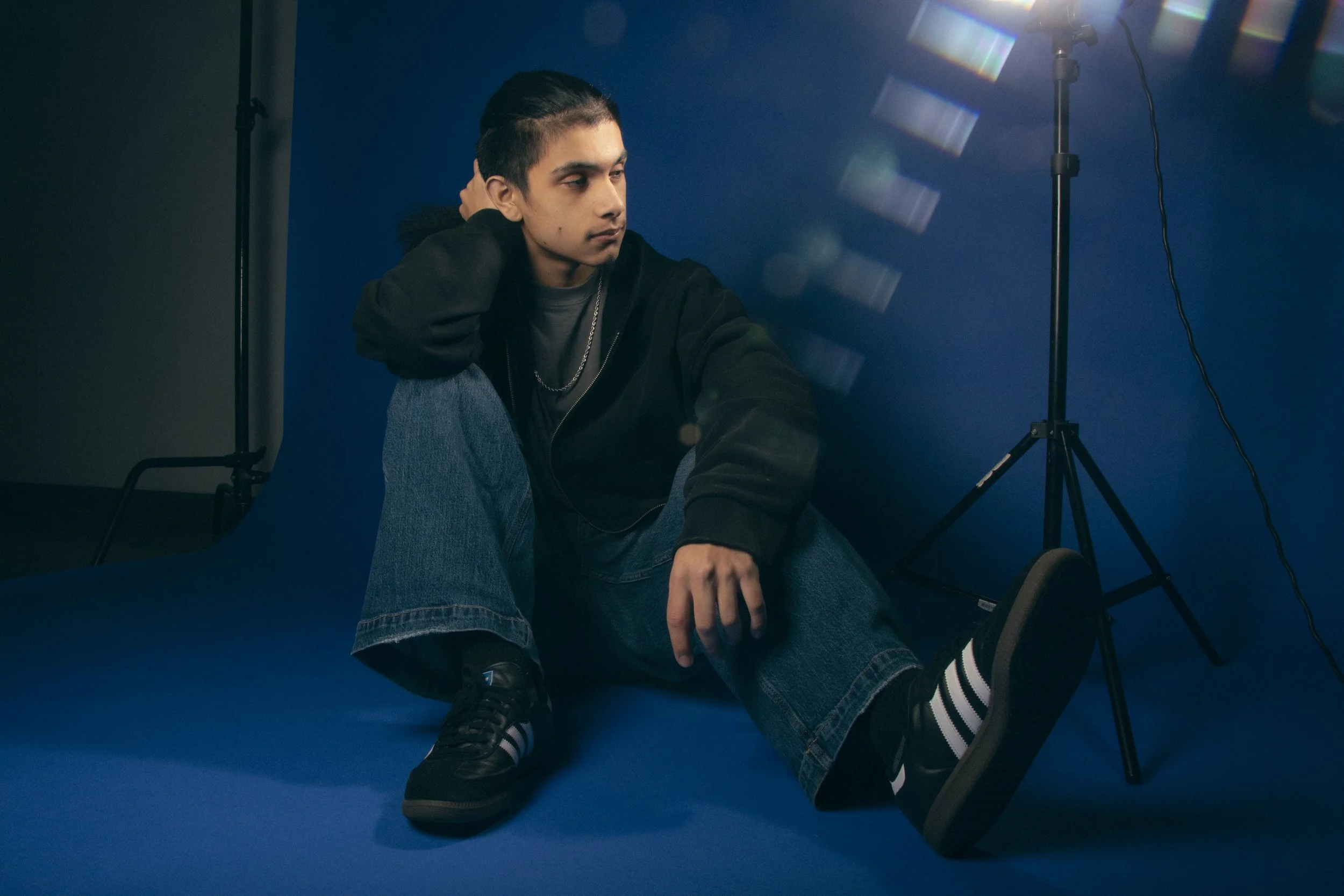 Young man with long hair tied back, sitting on a blue floor in front of a blue background, surrounded by professional photography lighting equipment, wearing black hoodie, gray t-shirt, blue jeans, and black sneakers with white stripes, with one hand