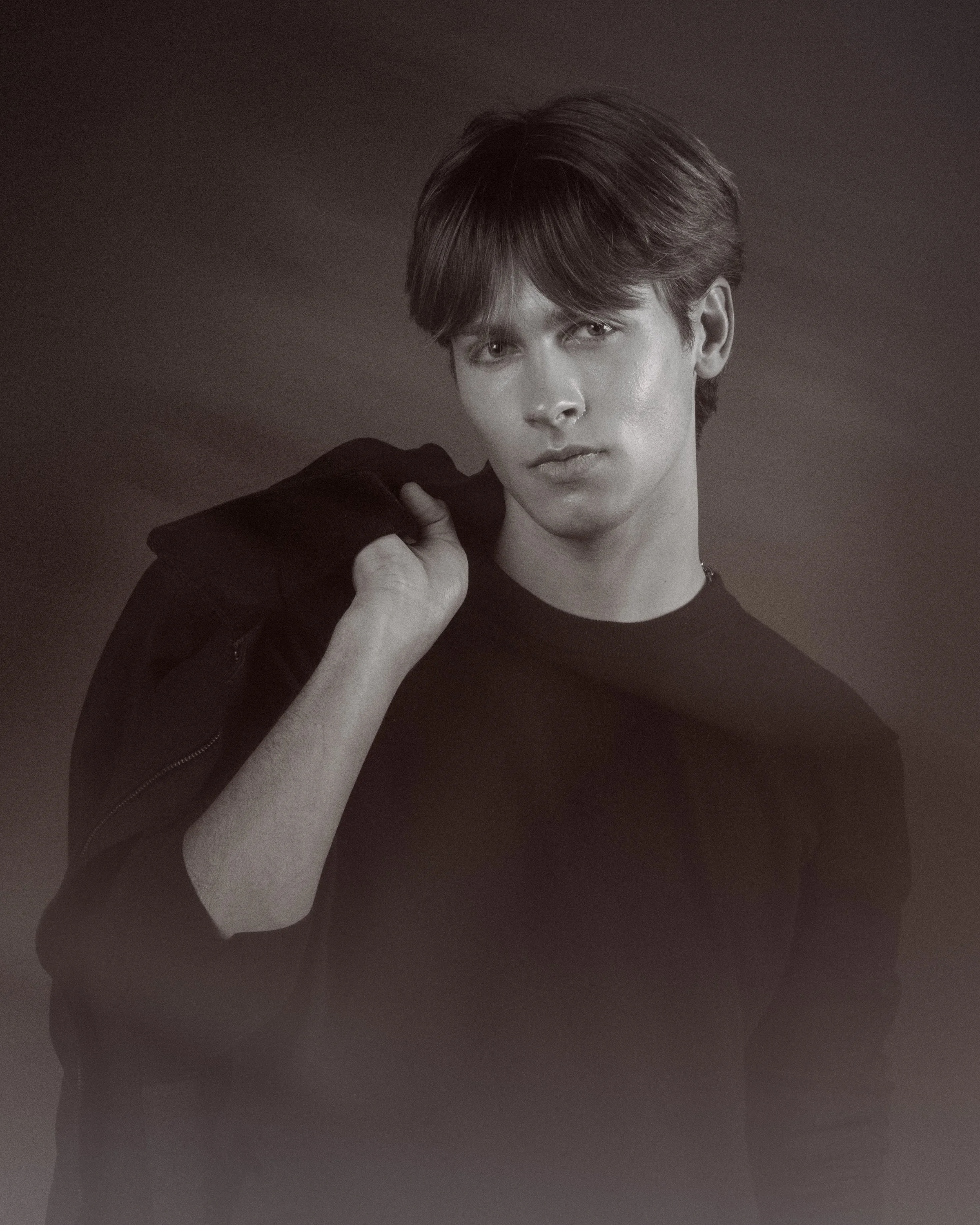Black-and-white portrait of a young man with medium-length hair, looking directly at the camera, with a serious expression on his face, wearing a dark jacket and holding the collar with one hand.