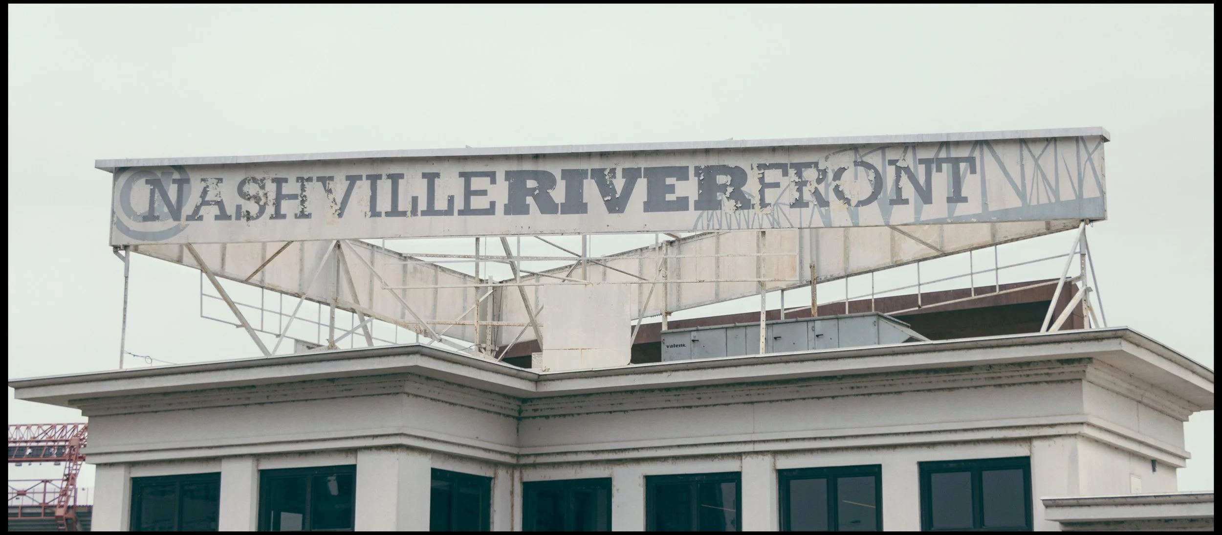 An aging, weathered sign on the rooftop of a building reads 'Nashville Riverfront' with the paint peeling off. The building has a white facade with large windows, and part of a crane is visible in the background.