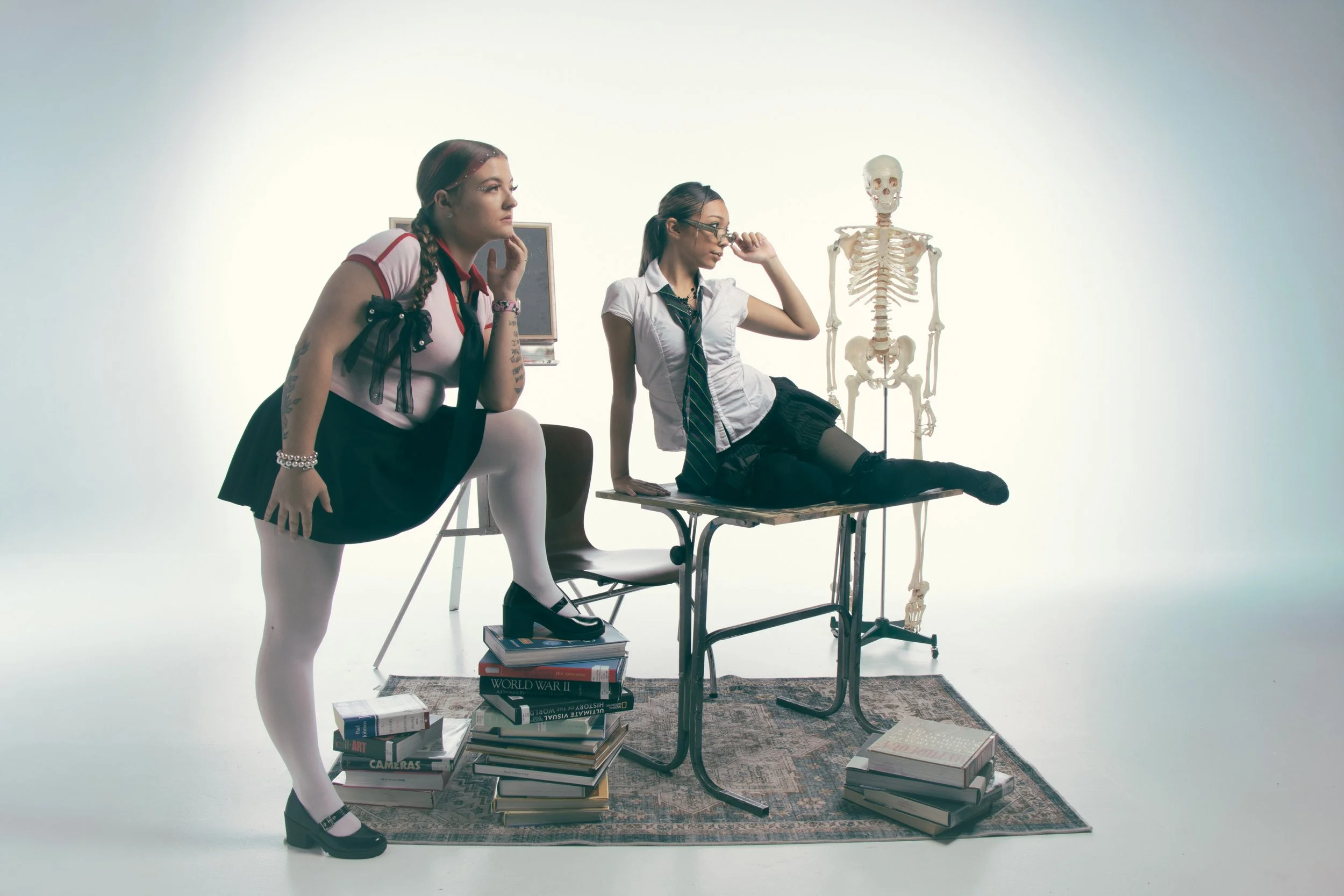 Two young women in school uniforms, one standing with her foot on a stack of books, the other sitting on a desk, next to a human skeleton model in a minimalist room with scattered books and a rug.