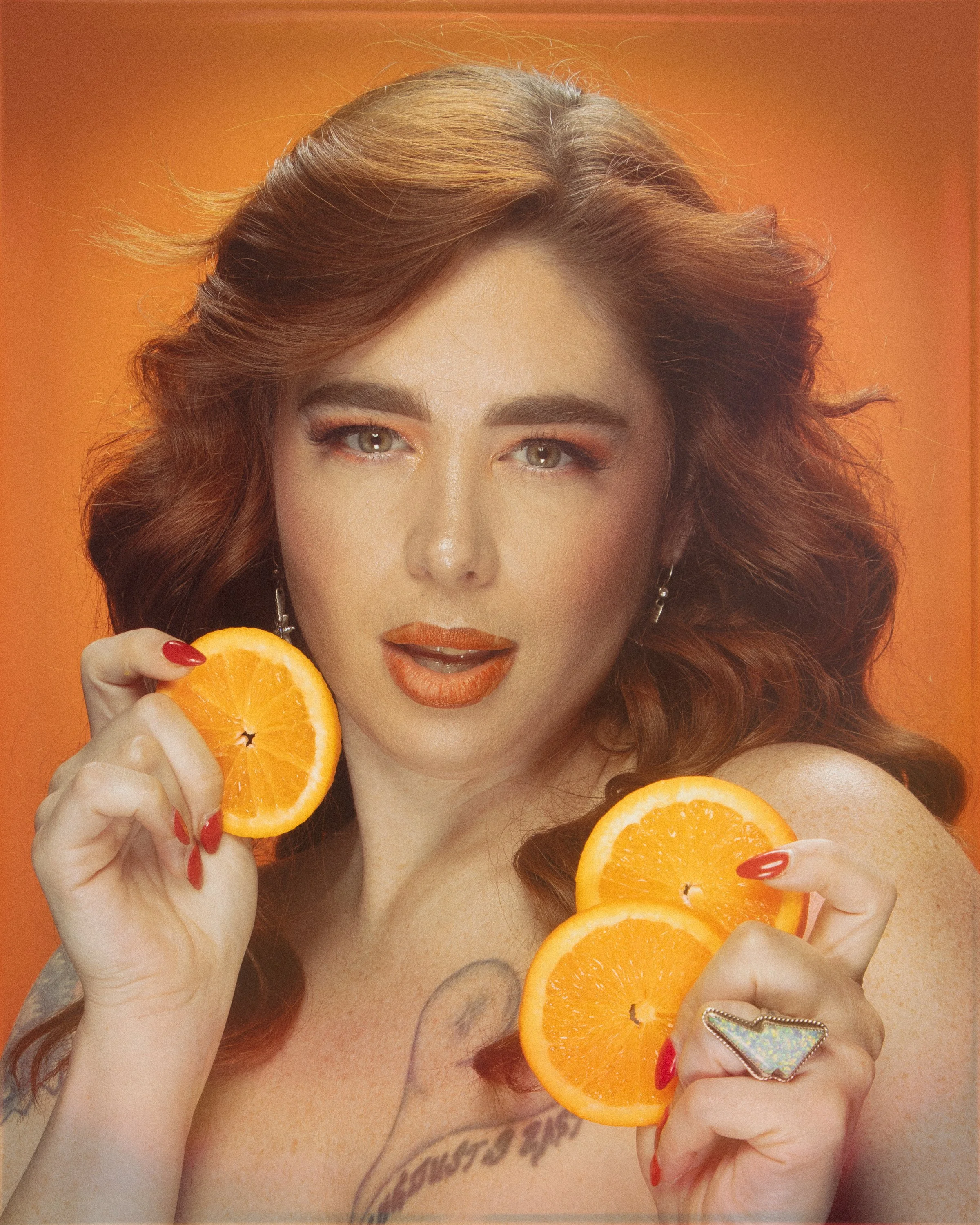 A woman with curly red hair and makeup holding orange slices near her face against a warm orange background.