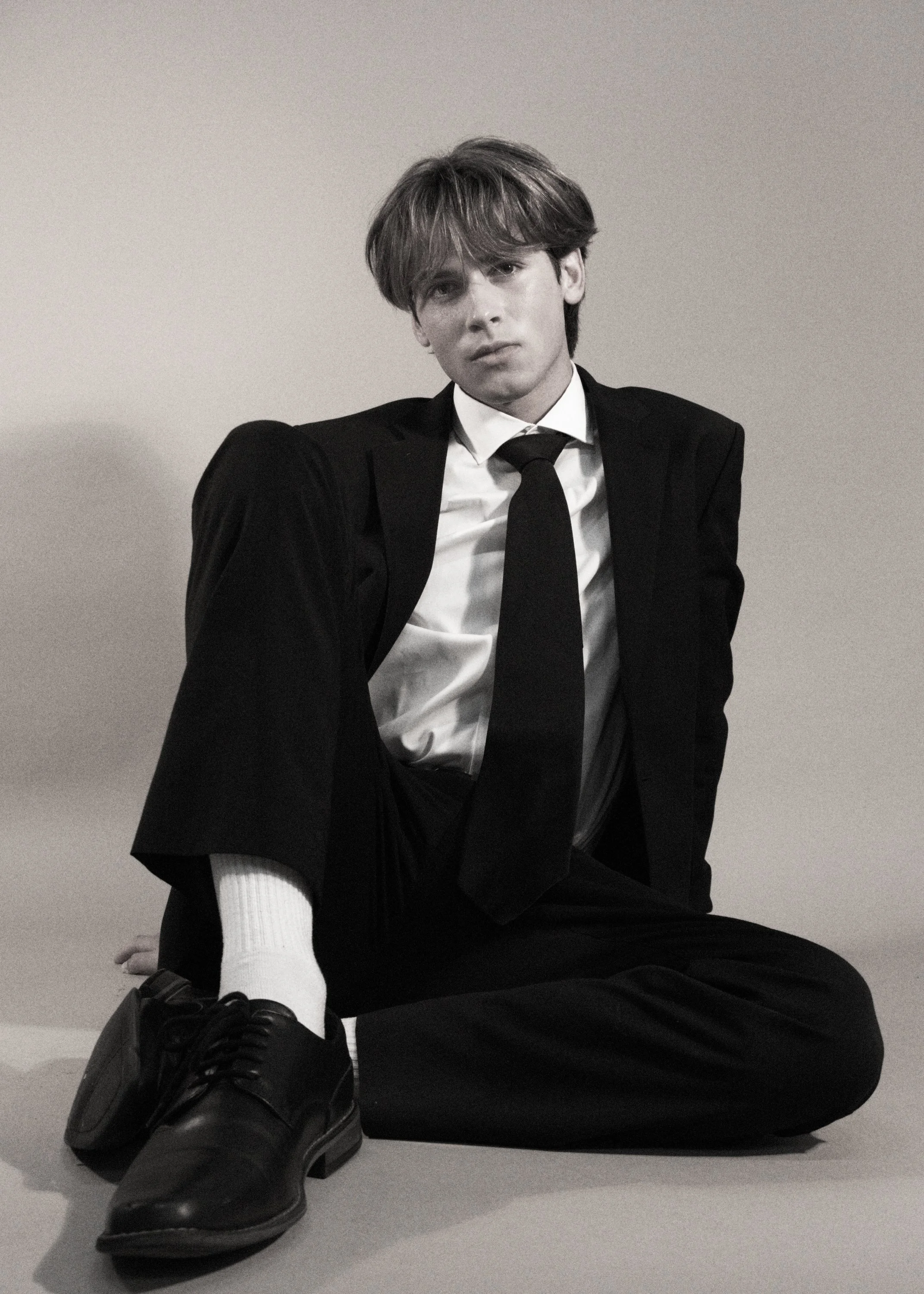 A black and white photo of a young man sitting on the floor against a plain wall. He is wearing a black suit, white shirt, black tie, black shoes, and white sock on one leg. His expression is neutral, and his hair is styled in a messy, wavy manner.
