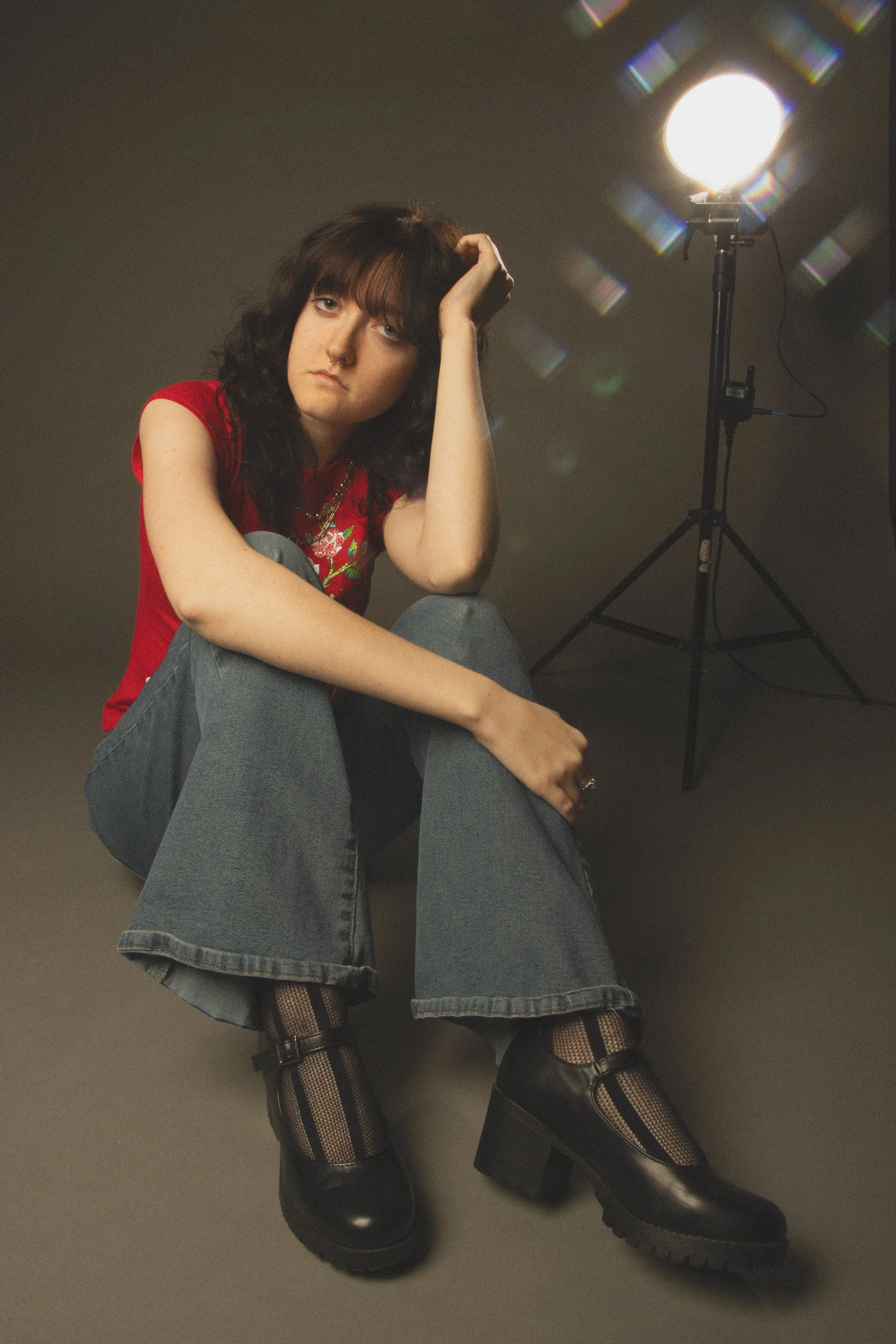 A person with dark curly hair wearing a red top, ripped jeans, fishnet stockings, and black platform boots sitting on the ground with one arm resting on their knee, in front of studio lighting.
