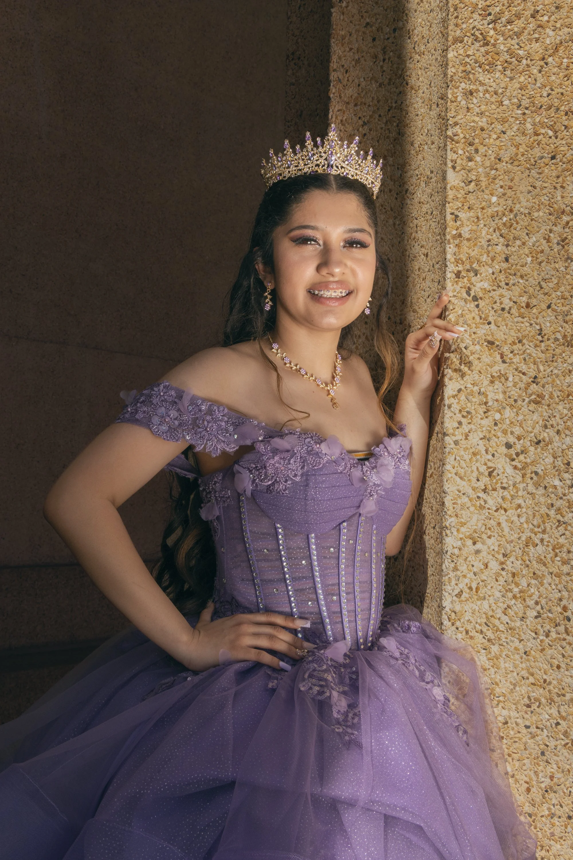 A young woman in a purple dress and jewelry, wearing a crown, smiling, standing against a textured wall.