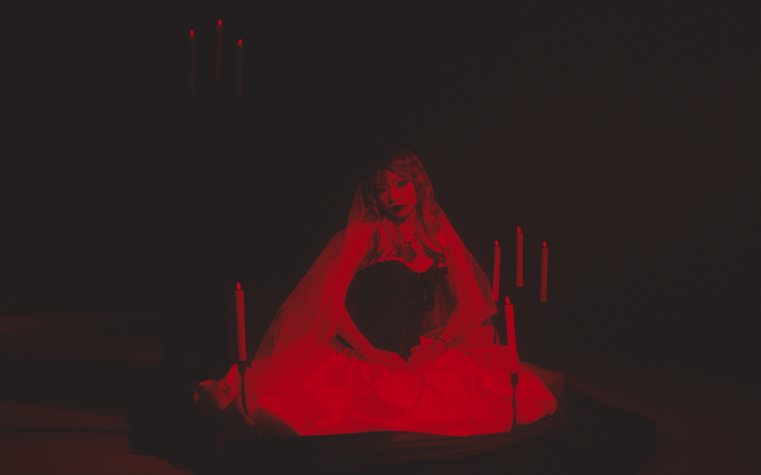 A woman dressed in black with a sheer red cape sitting on the ground surrounded by tall red candles, in a dark setting.