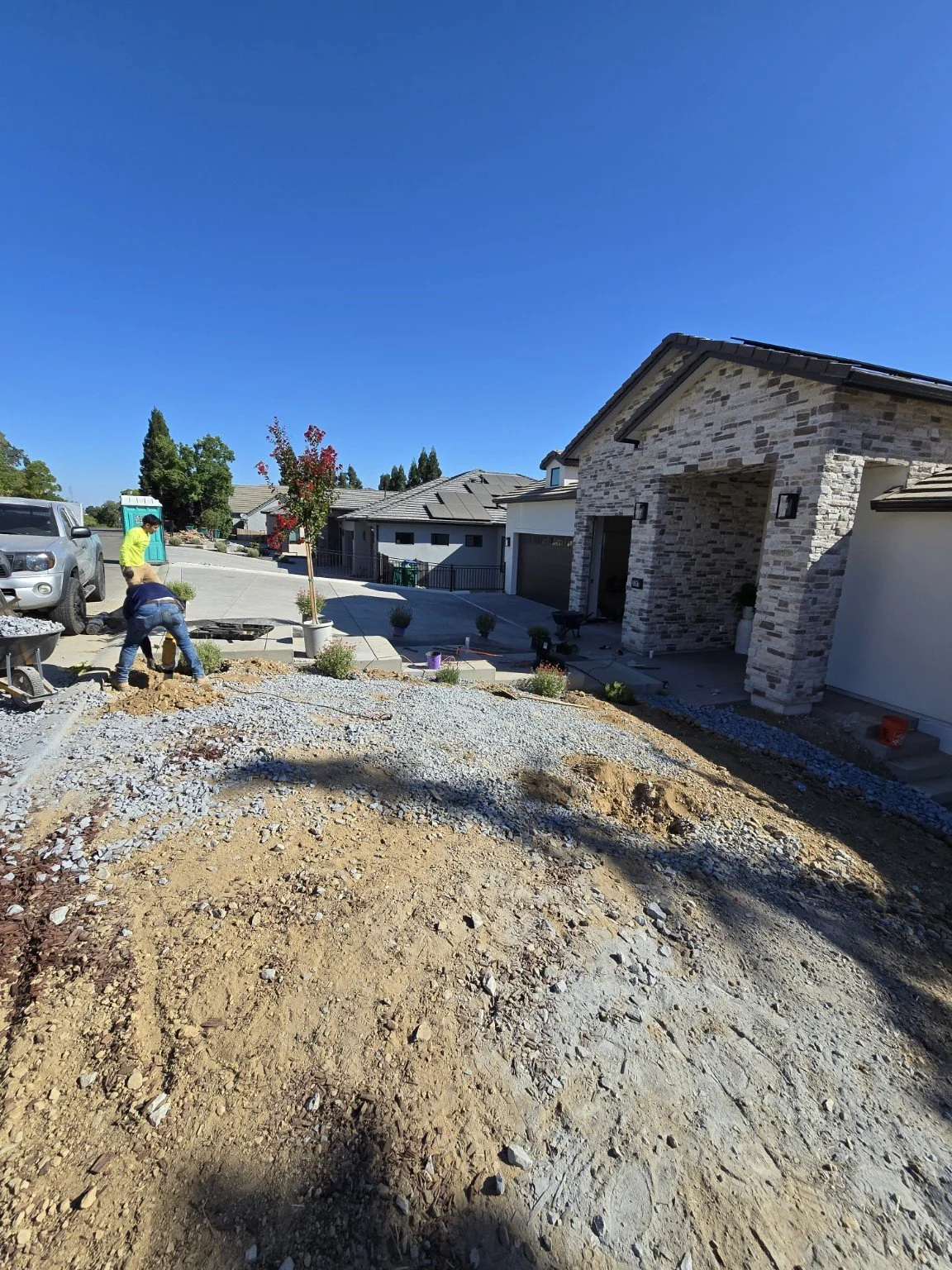 Construction workers working on a driveway in front of a house with brick facade, with a car parked on the street and a blue portable toilet in the background.