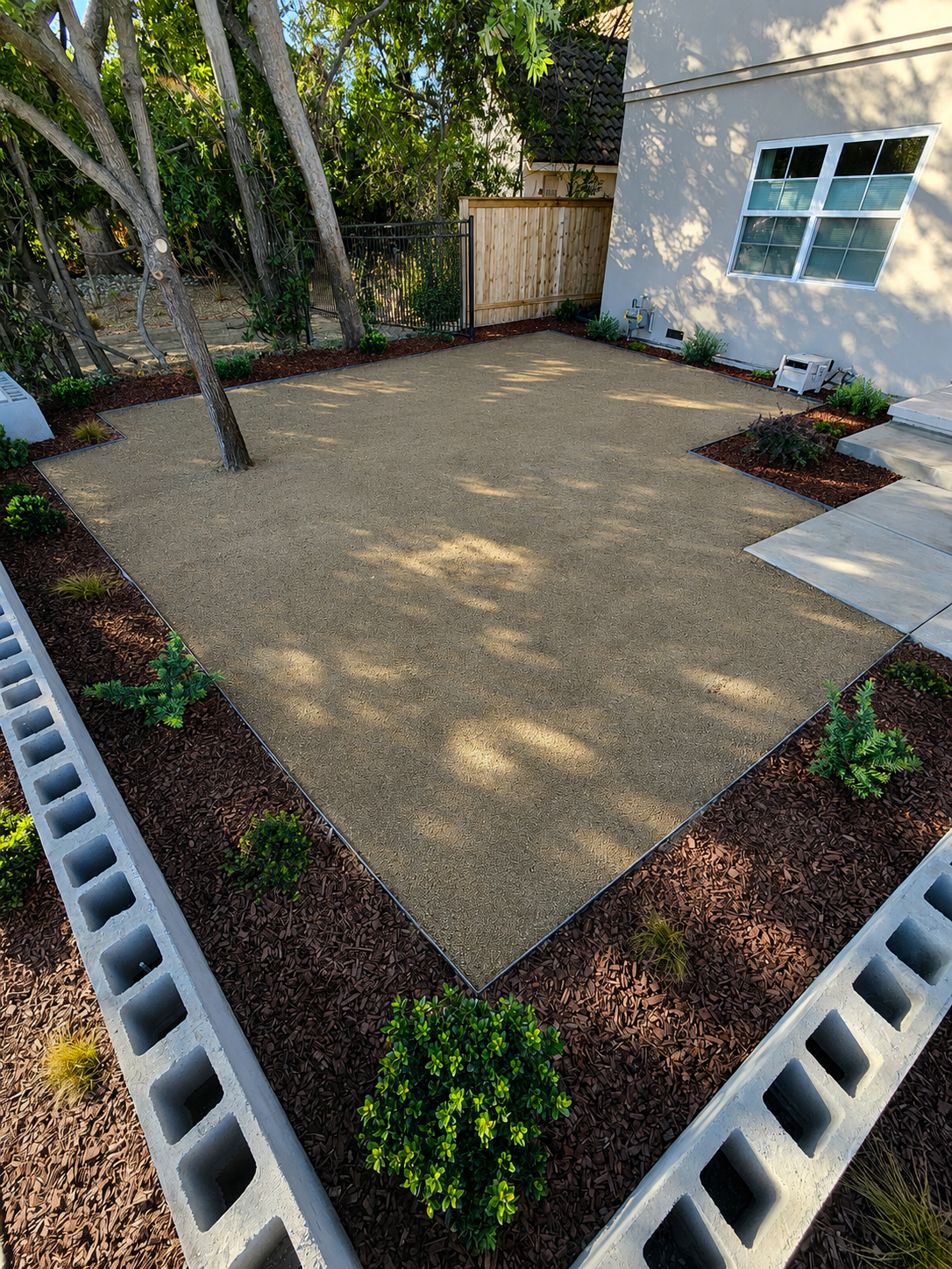 Backyard with a level sandy area, surrounded by garden beds with small plants and mulch, bordered by cinder blocks, and a white house with a window and steps on one side.