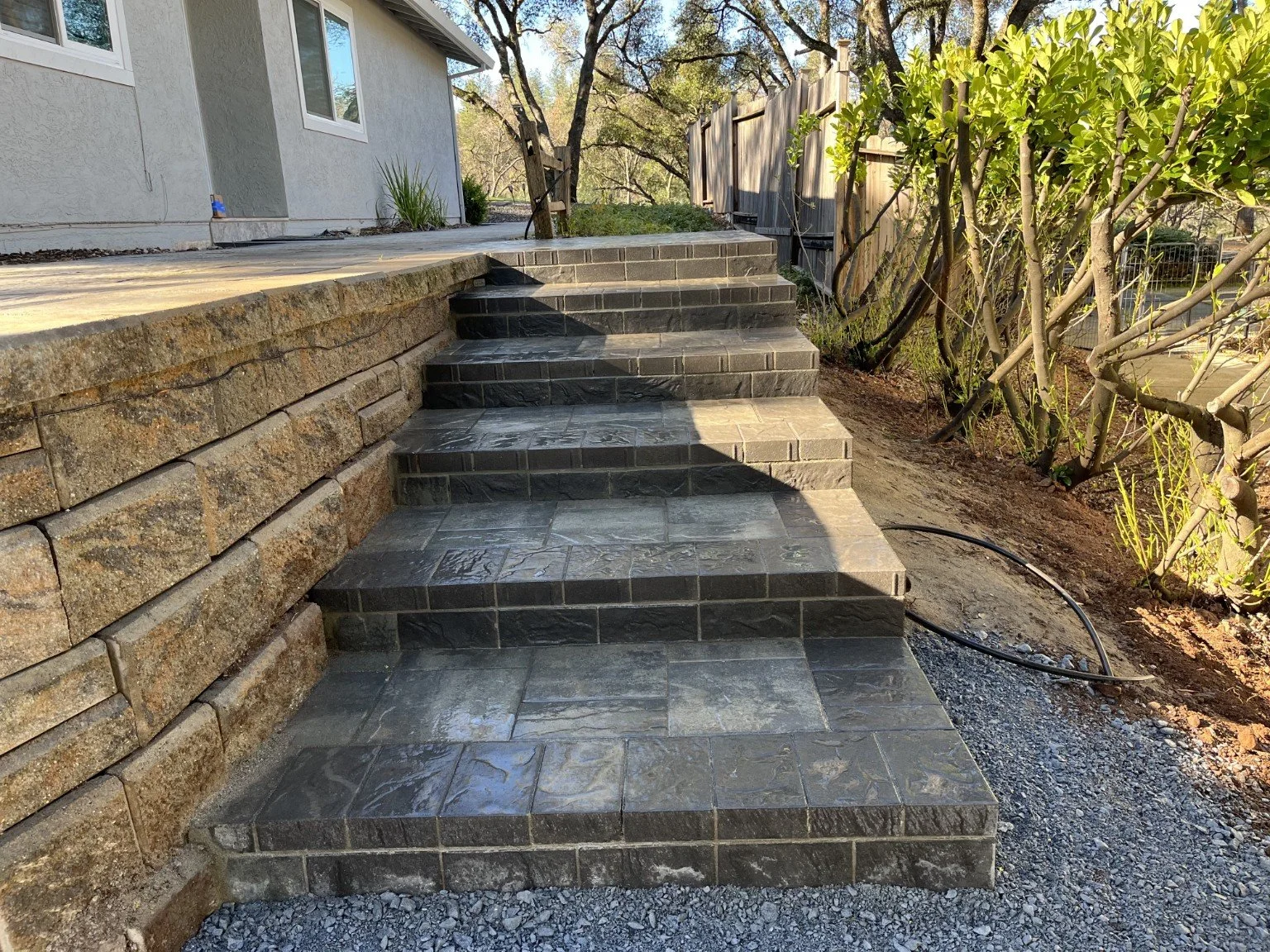 Stone steps leading up to a house with a gray exterior and white window frames, surrounded by greenery and wooden fencing.