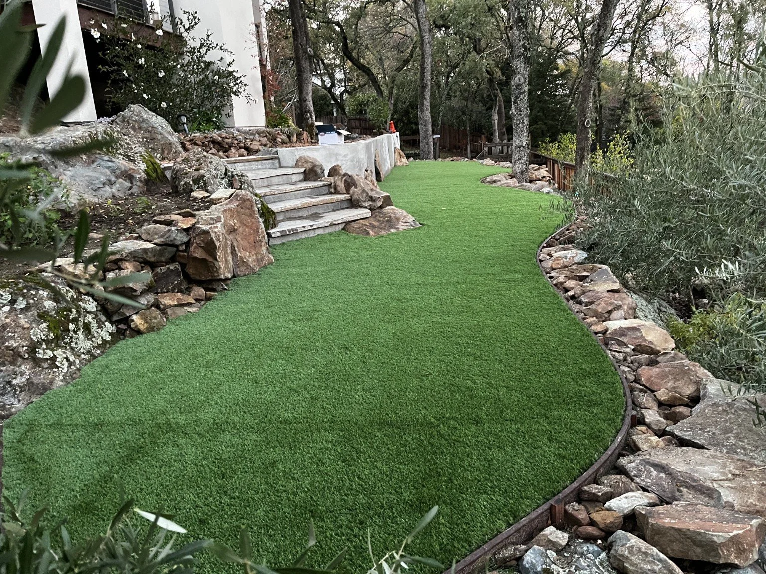 A landscaped backyard with artificial grass, stone steps leading up to a patio, and a surrounding rock border with trees and bushes.