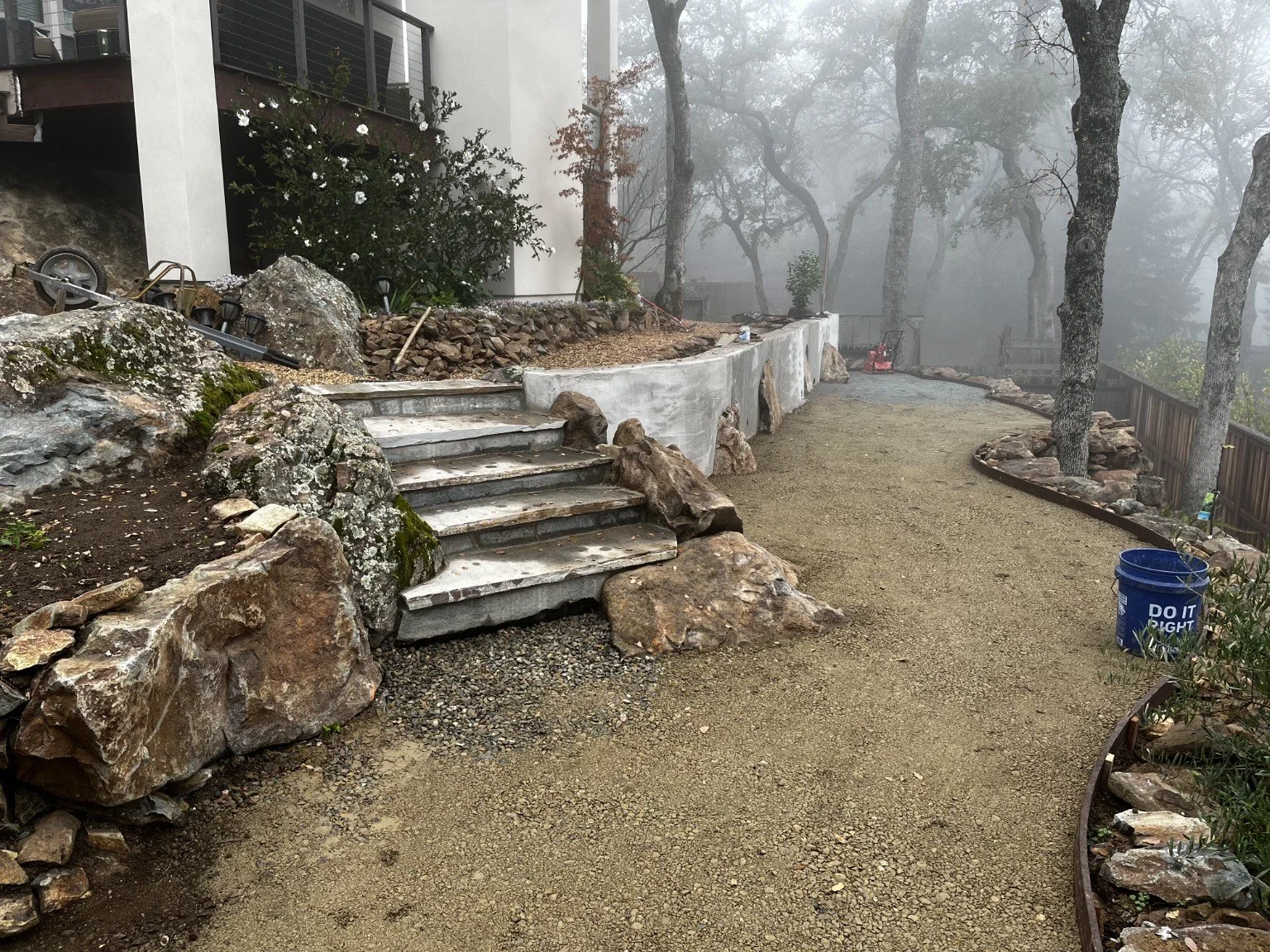 A foggy outdoor scene with a gravel pathway under construction, bordered by a wooden fence and trees, with stacked rock landscaping and construction tools nearby.
