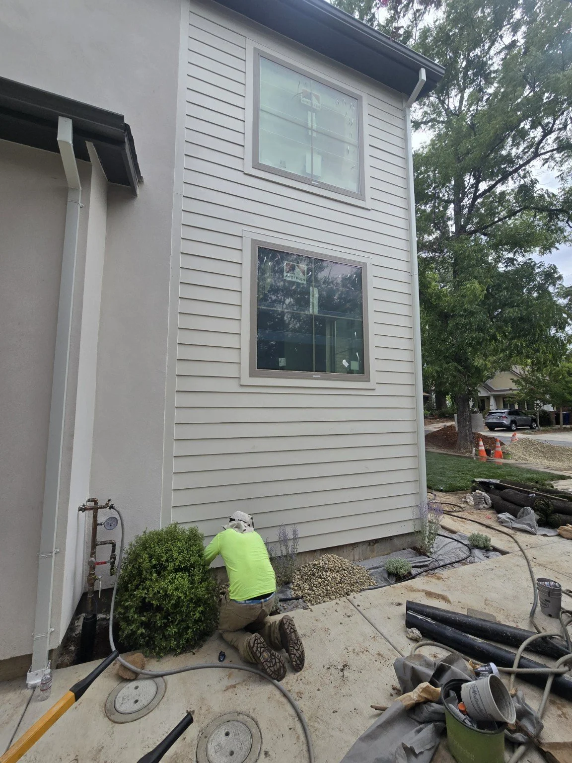 A construction worker kneeling on the ground near a small green bush in front of a house under construction. The house has beige siding and large windows, with construction materials and tools scattered around.