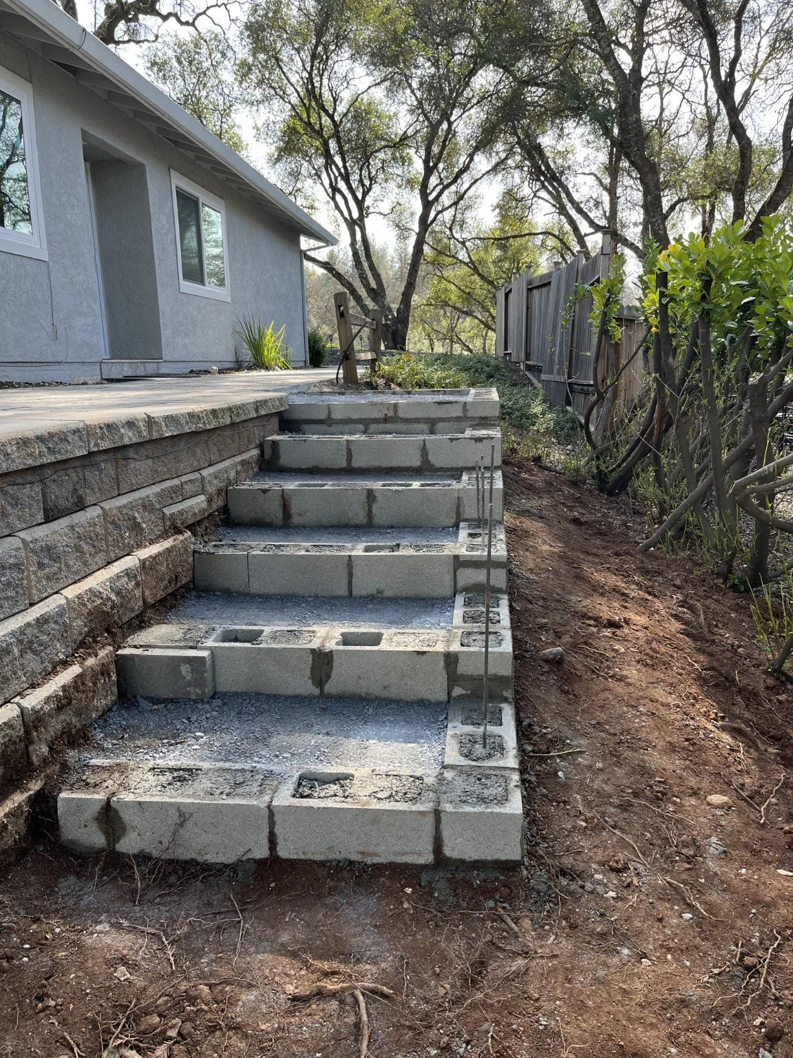 Unfinished concrete staircase construction in backyard with dirt pathway on the right, wooden fence, and house with gray exterior on the left. Trees in background.