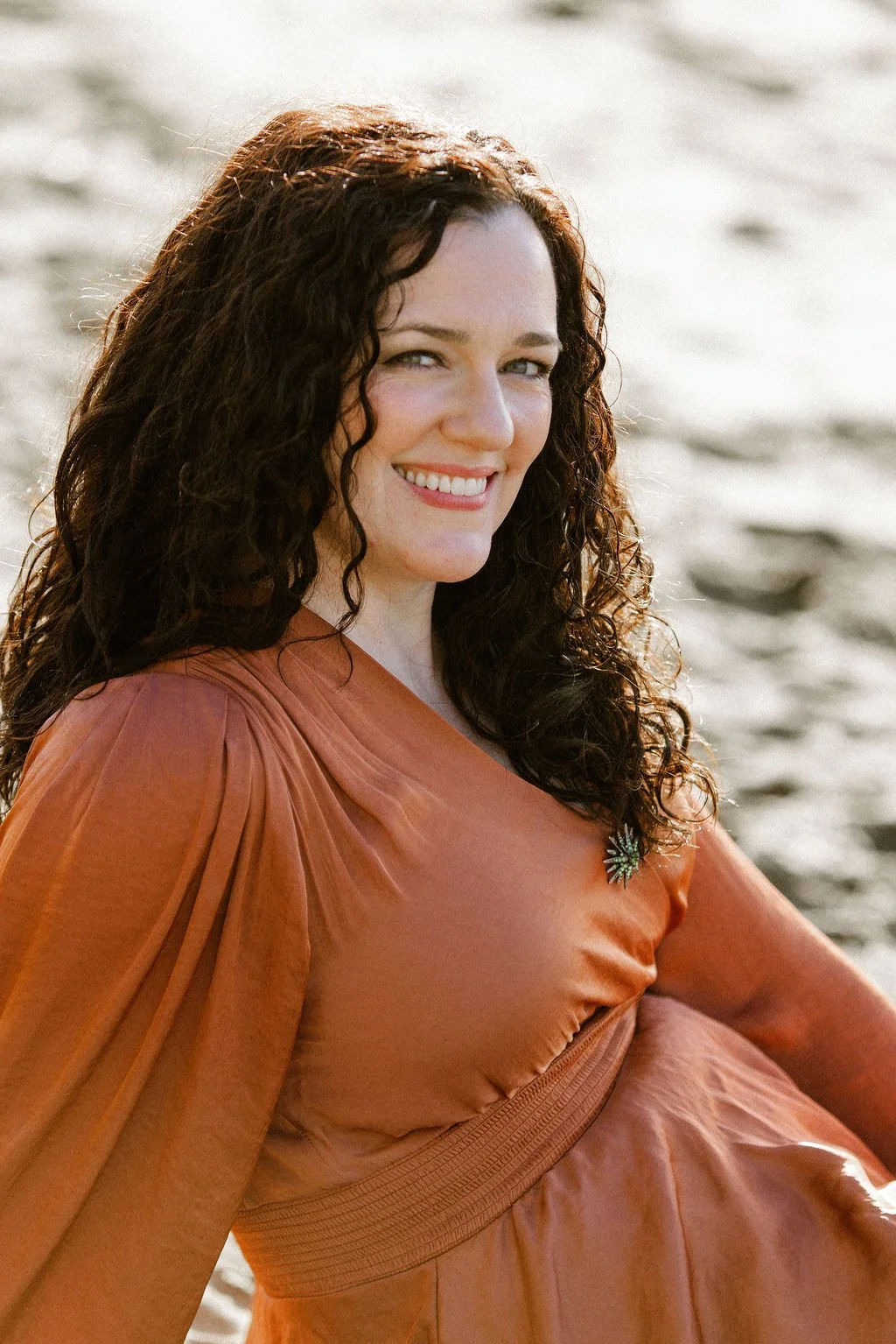 Lindsay Lackey, a woman with curly brown hair smiling outdoors in warm sunlight, wearing an orange dress with a decorative brooch on her chest, sitting on sandy ground.