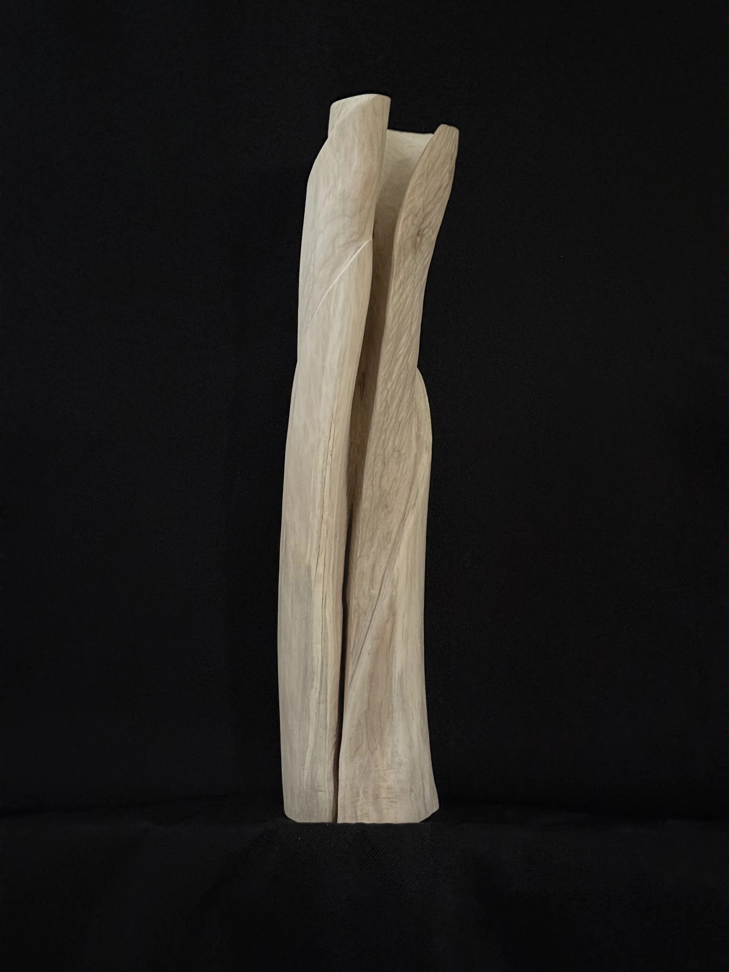 Full-length view of an elongated, hand-carved modern wood sculpture with an intertwined split form against a studio black backdrop.