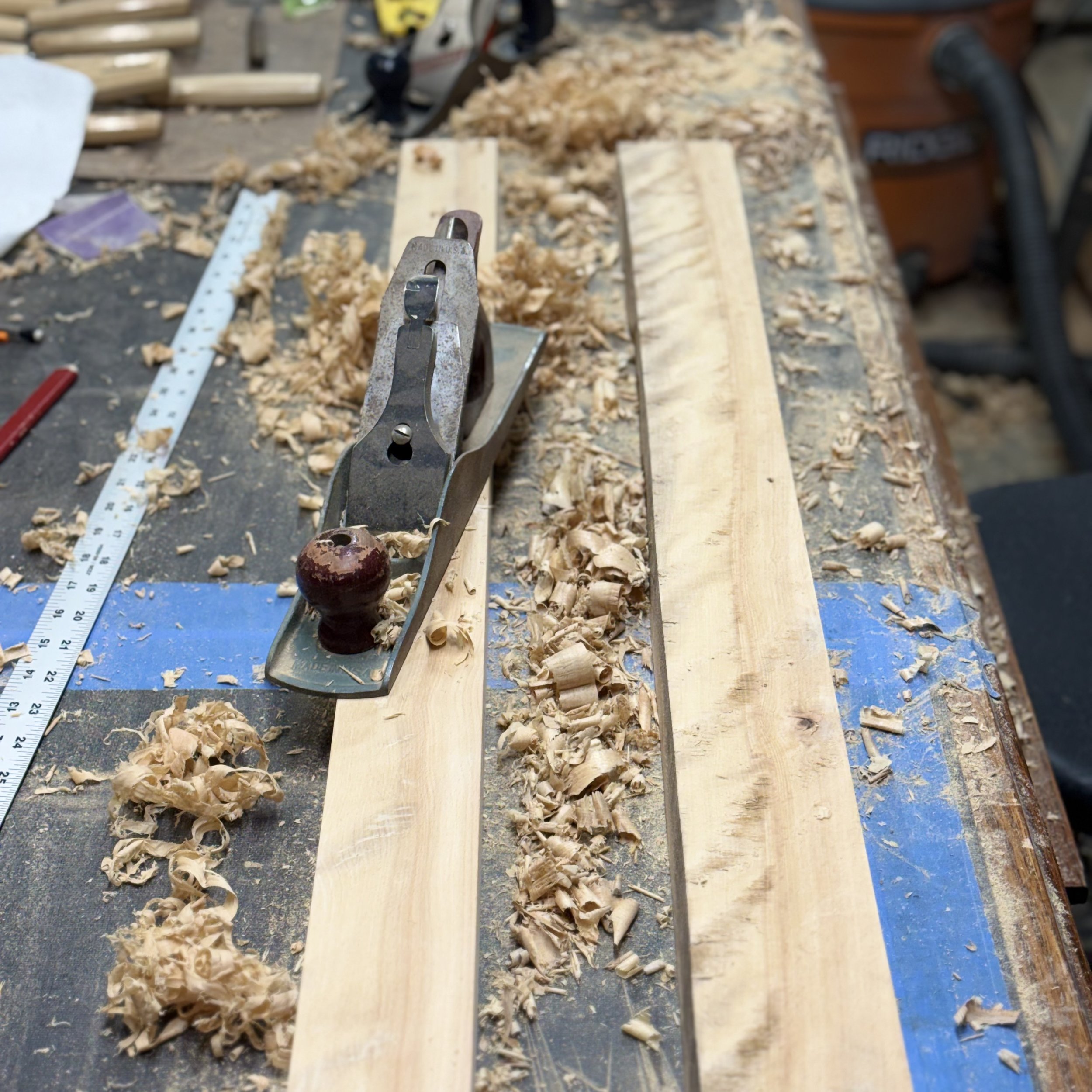 A woodworking workbench with a hand planer, a metal ruler, and shavings from wood surfaces. A large piece of wood is clamped in place, and there is a power tool handle visible on the right.