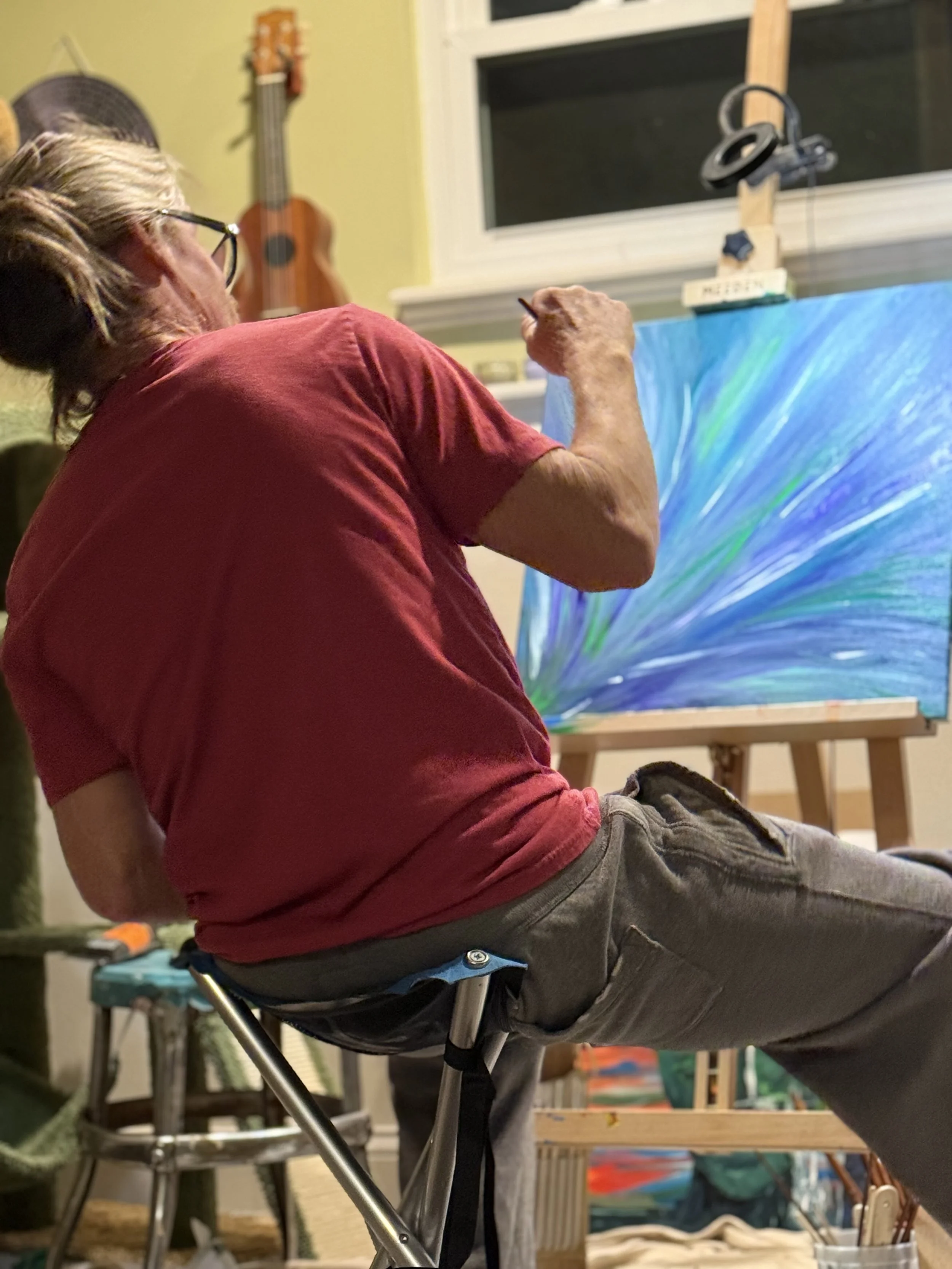 A person with long hair is sitting on a chair, painting on a canvas. The scene appears to be a home art studio with musical instruments and art supplies in the background.