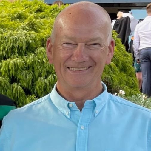 Smiling middle-aged man with a bald head wearing a light blue button-up shirt outdoors against green foliage and people in the background.