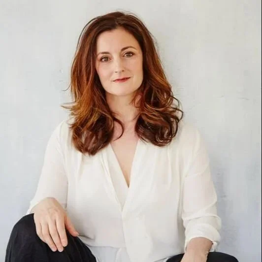 A woman with long wavy auburn hair, wearing a white blouse, sitting with her hand resting on her knee, against a plain light background.
