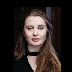 Portrait of a young woman with long brown hair and a black top standing indoors against a dark background.