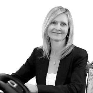 Black-and-white photo of a woman with shoulder-length blonde hair, wearing a blazer and white top, smiling and seated at a desk.