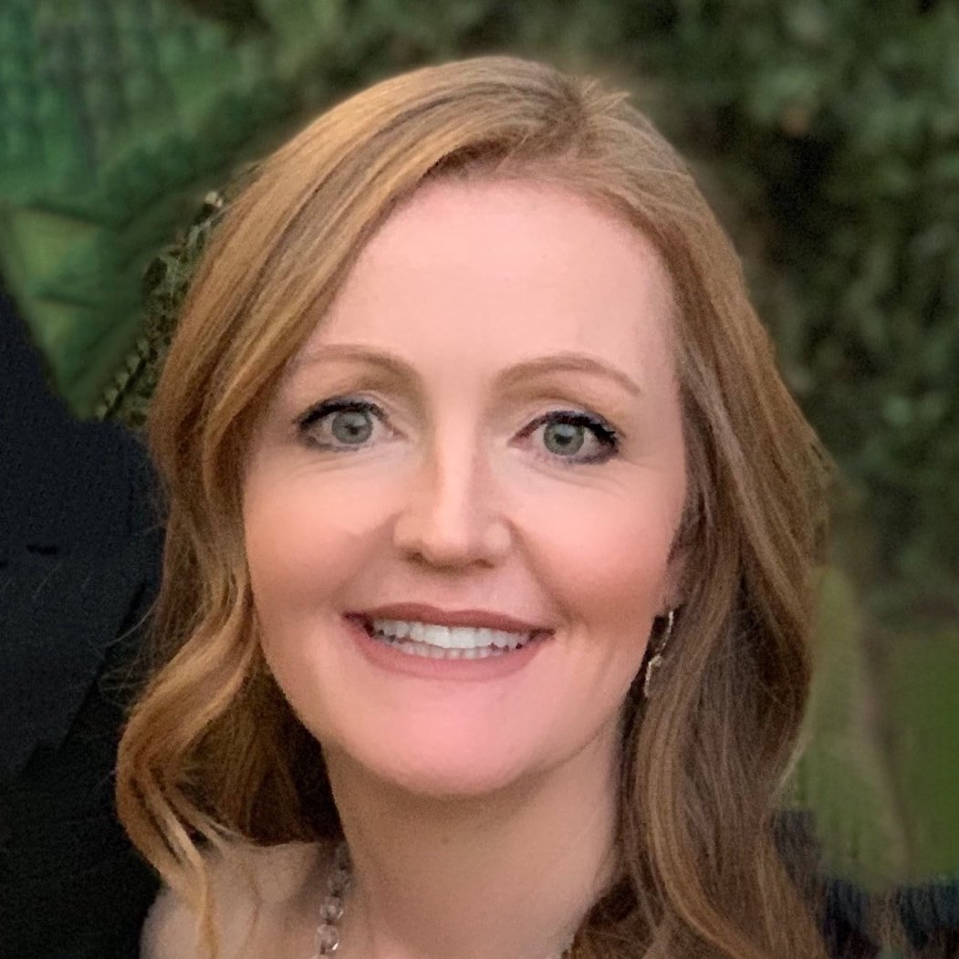 A woman with red hair smiling outdoors with greenery in the background.