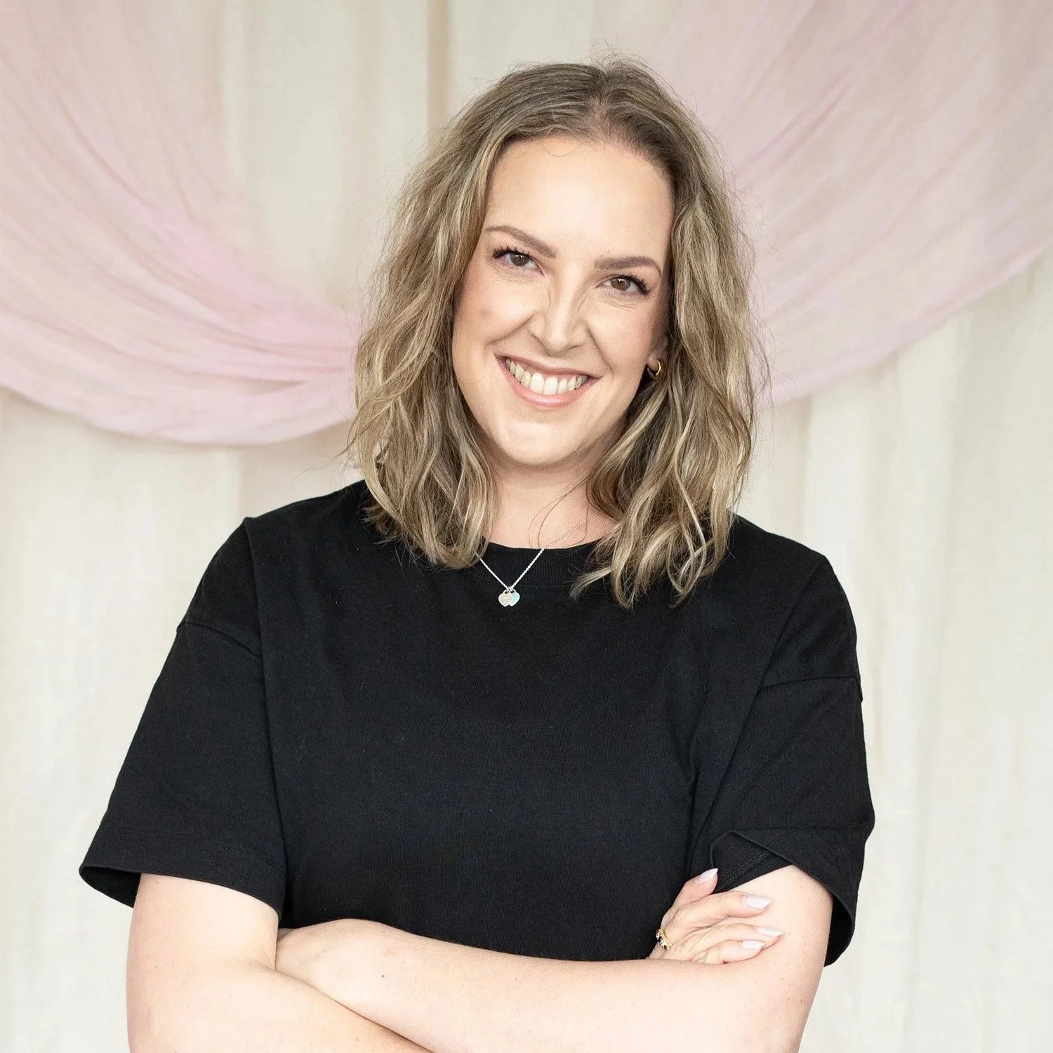 A woman with shoulder-length wavy blonde hair, wearing a black top, smiling, standing with arms crossed in front of a light-colored background with pink and purple abstract art.