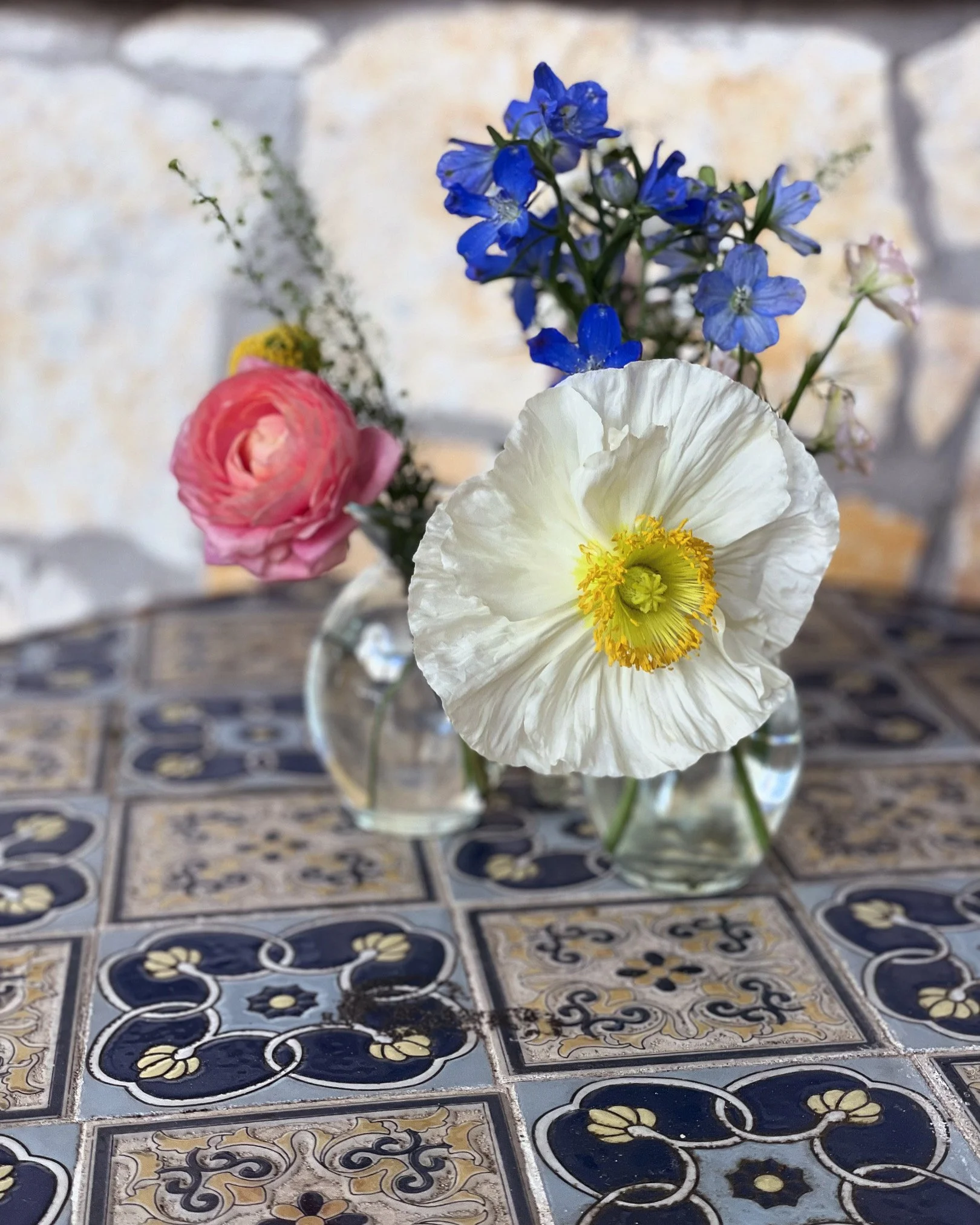 A small bouquet of fresh flowers in a clear glass vase on a decorative tiled surface. The bouquet contains white, pink, and blue flowers.