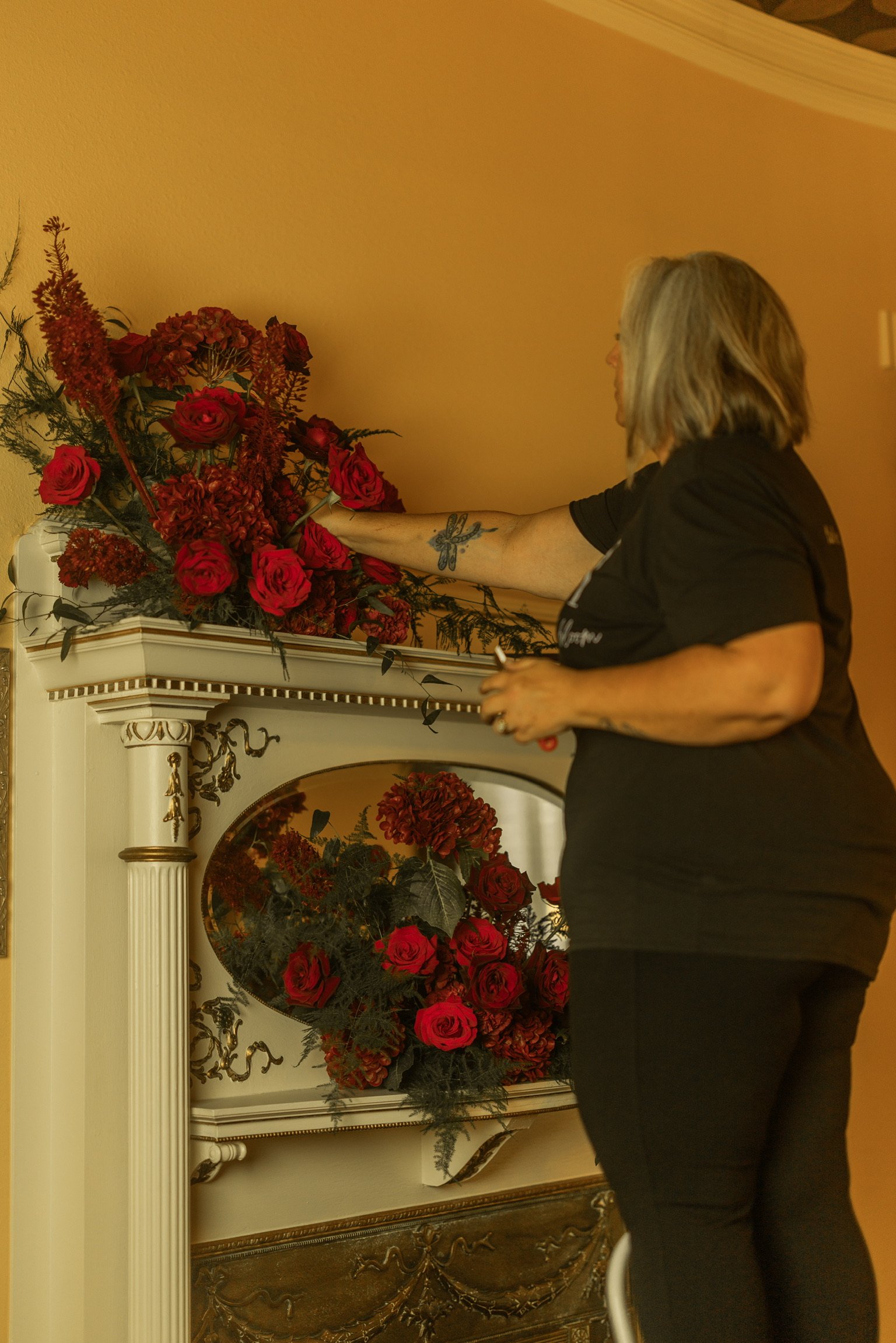 A floral designer with gray hair arranging red roses in a floral display on a white ornate fireplace mantel.