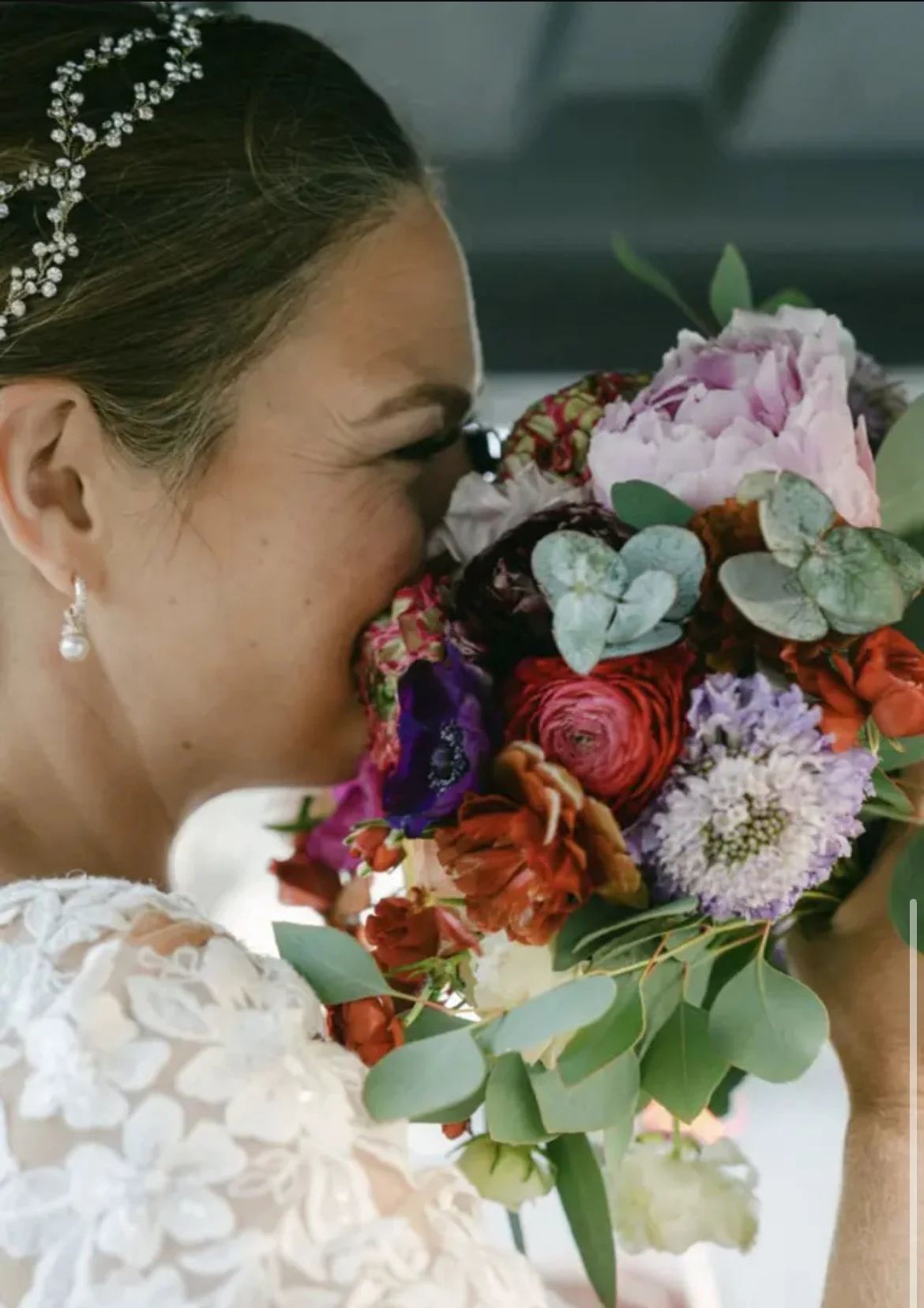 Close-up of a bride with dark hair, wearing pearl earrings and a white lace dress, smelling a colorful bouquet of flowers including pink, purple, red, and green leaves.