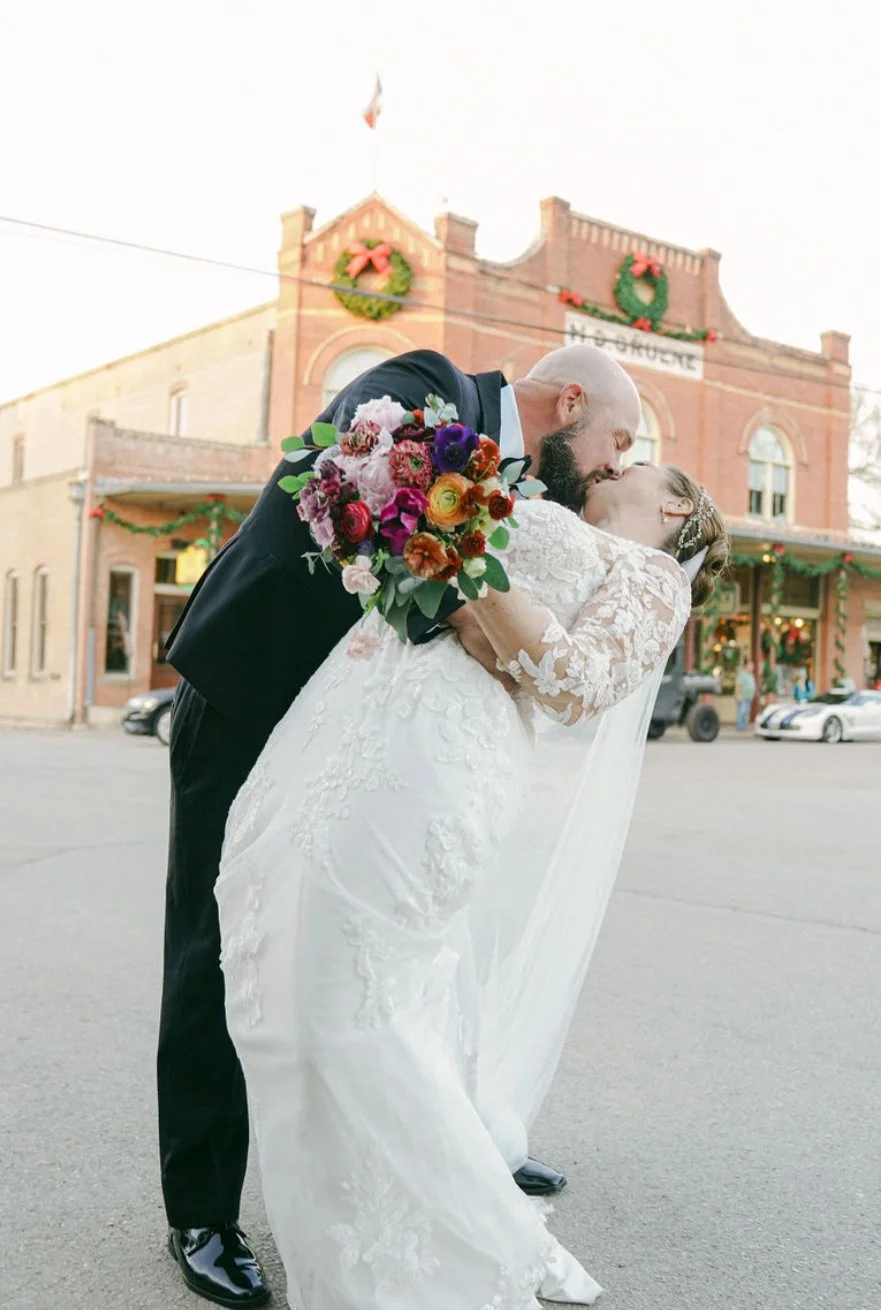 A bride and groom share a kiss on a street, with the groom dipping the bride. The groom is holding a bouquet of colorful flowers. The background features a historic brick building decorated with holiday wreaths and garlands.