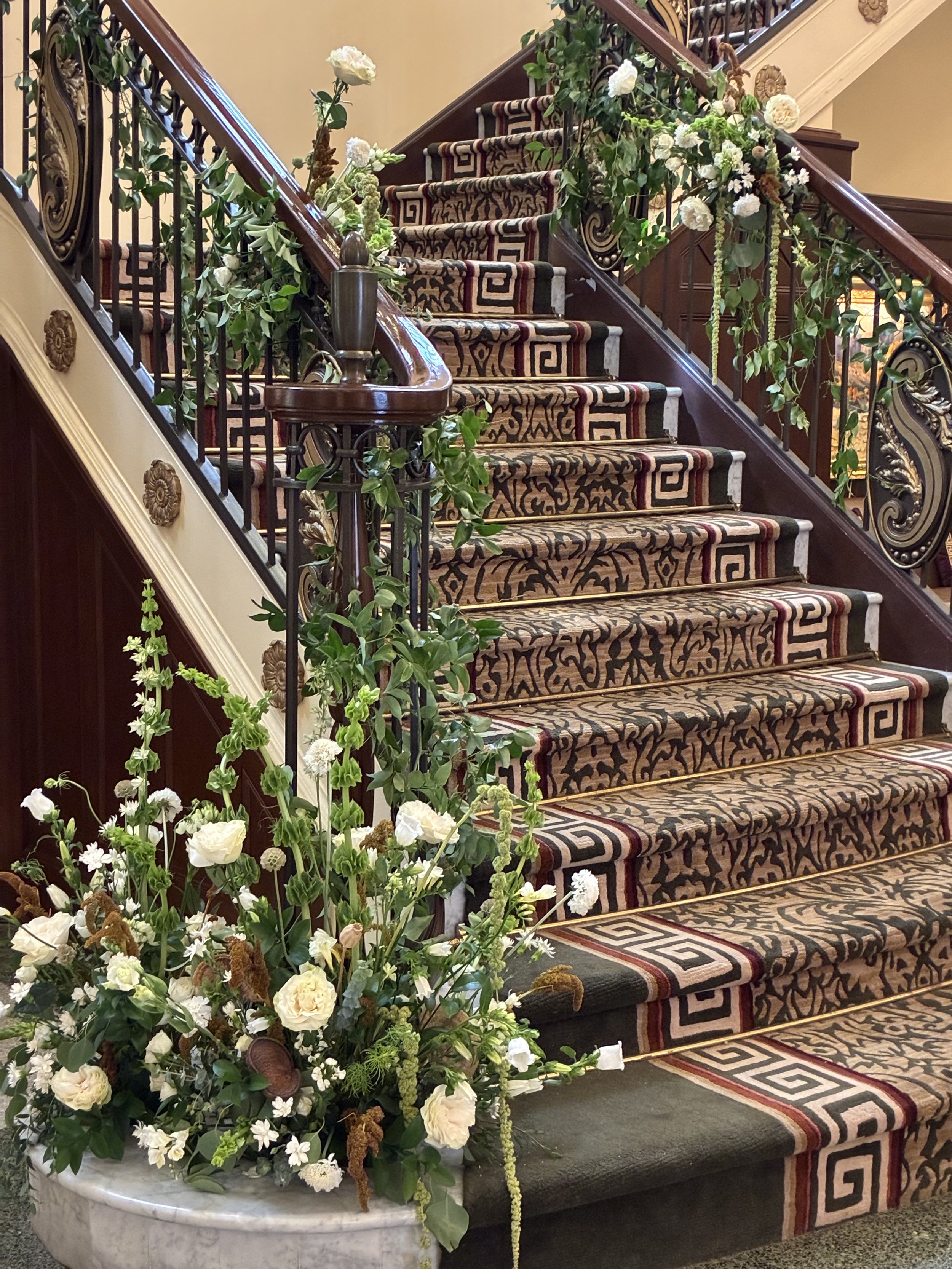 A grand staircase decorated with floral arrangements, with intricate patterned carpet and ornate wrought iron railings.