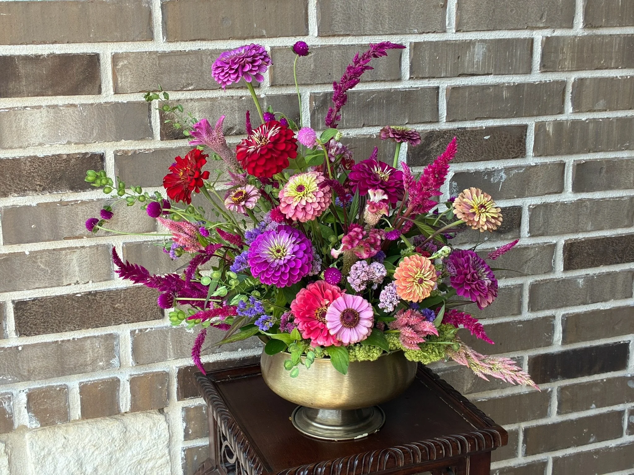 A colorful bouquet of various flowers in a brass vase set on a dark wood table against a brick wall background.