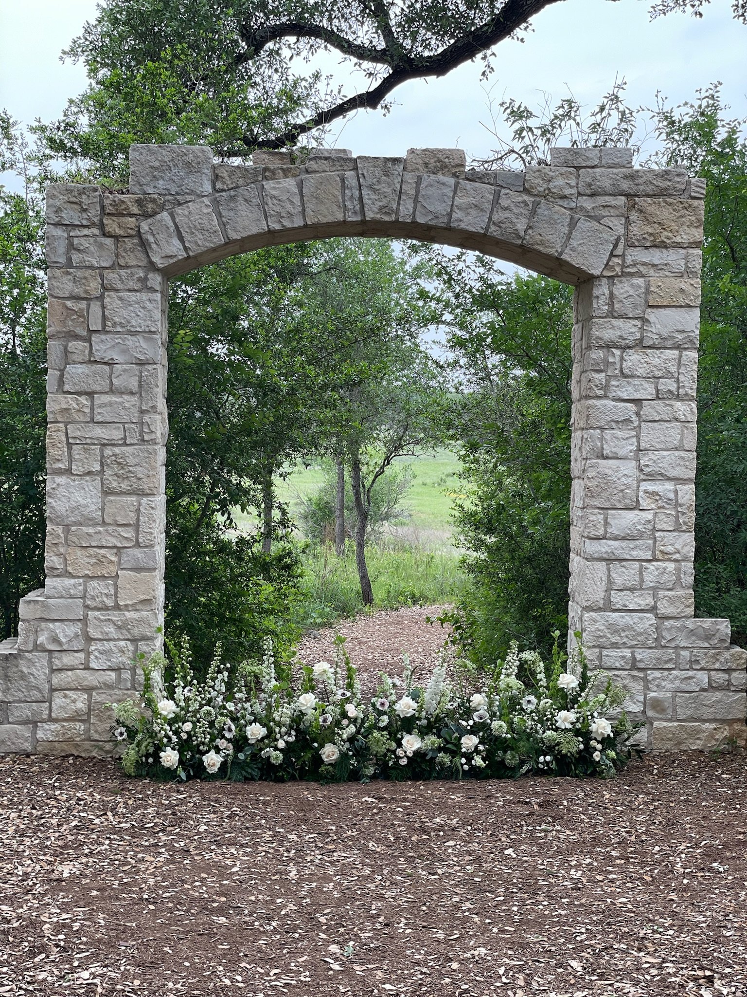 Stone archway with white flowers at the base, surrounded by green trees and a pathway leading through the arch to a grassy area beyond.