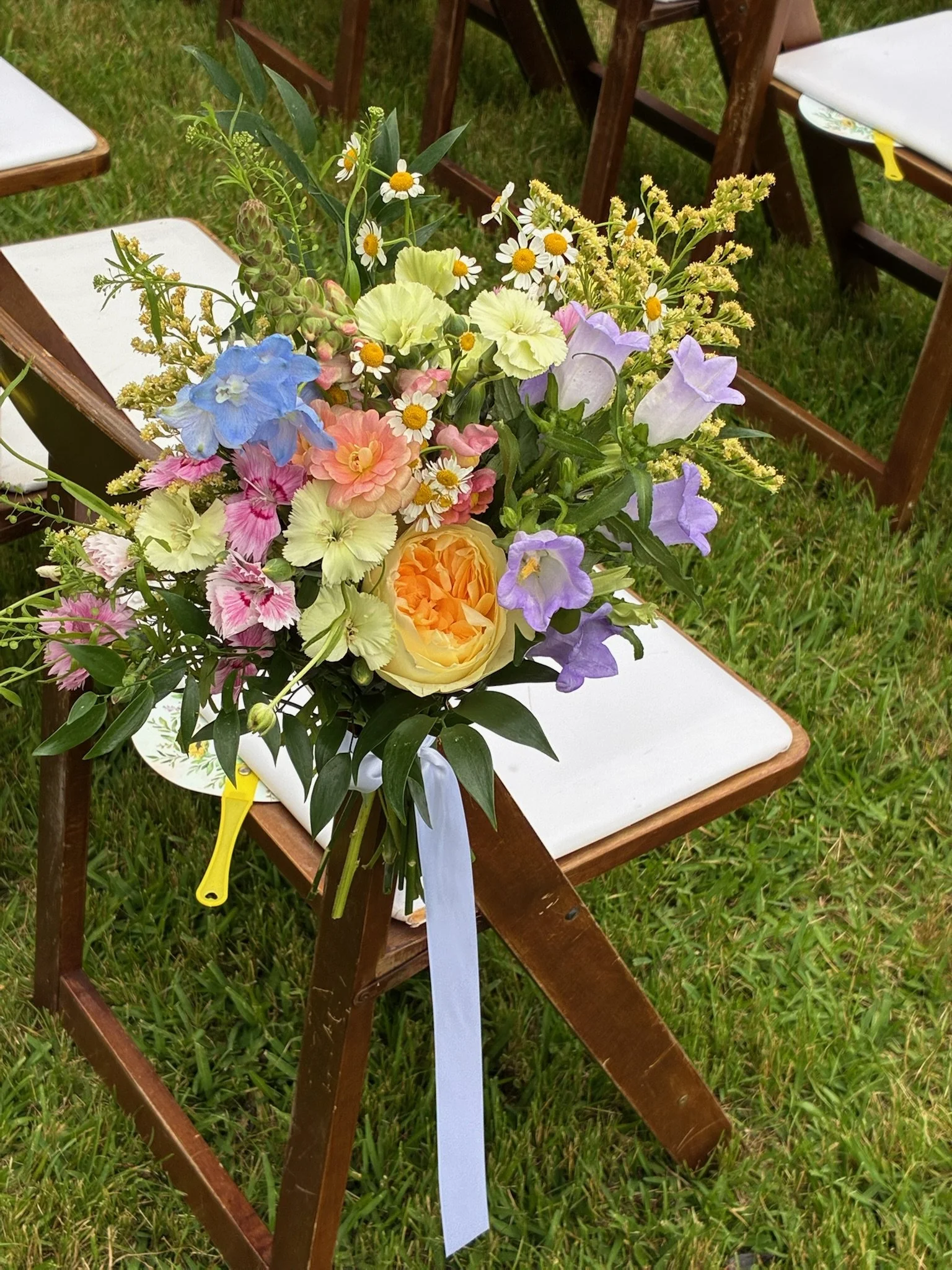 A floral bouquet placed on a wooden outdoor chair with white cushions, set on green grass. The bouquet includes daisies, lilies, roses, and other mixed flowers with green foliage, tied with a white ribbon and a yellow attached knife.