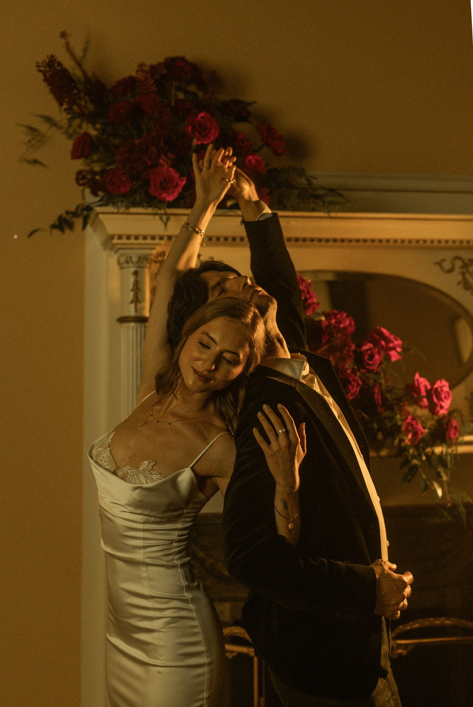A couple dancing closely in a warmly lit room with pink and red roses in the background.