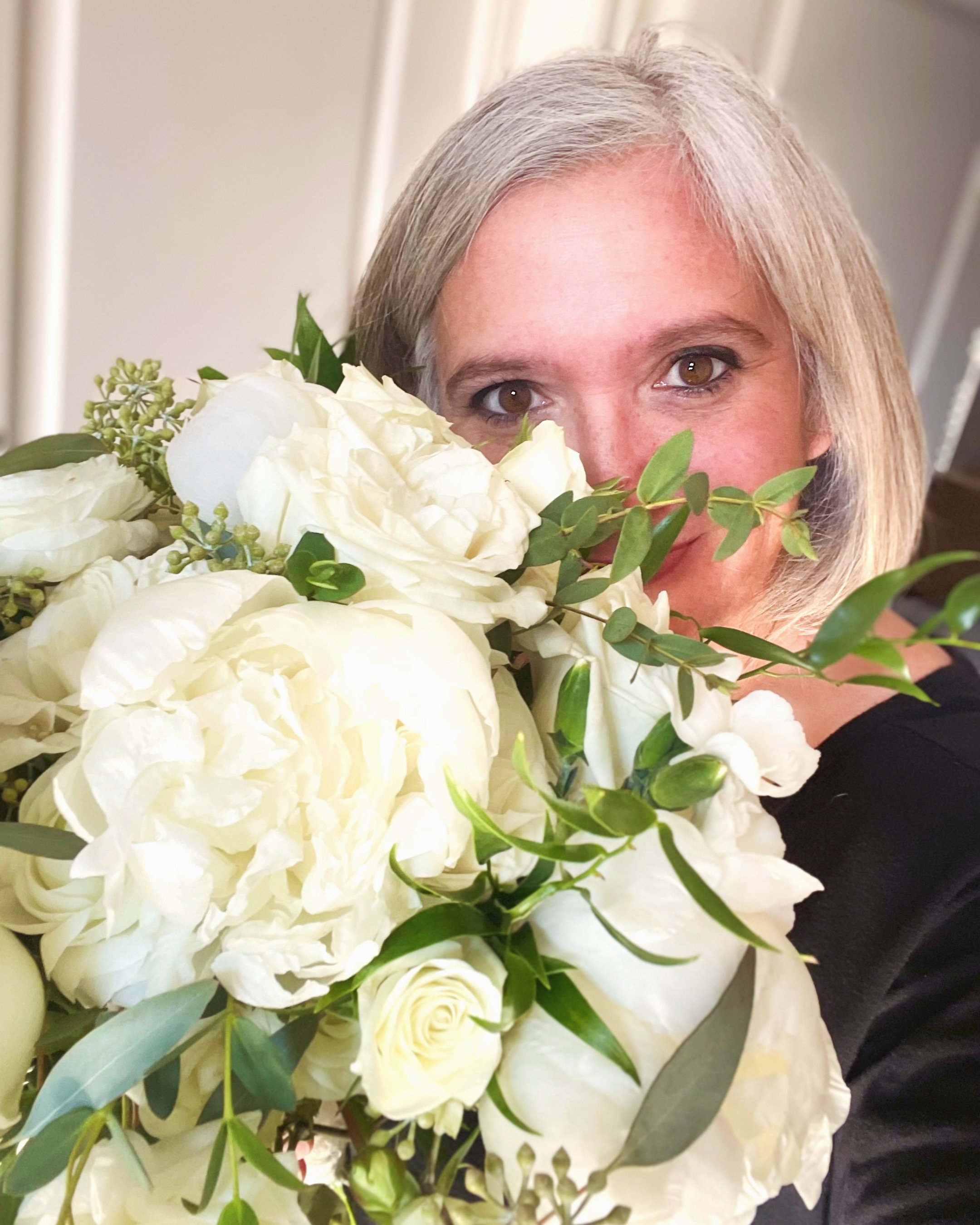 A woman with gray hair holding a large bouquet of white flowers, including roses and other blooms, partially covering her face as she smiles.
