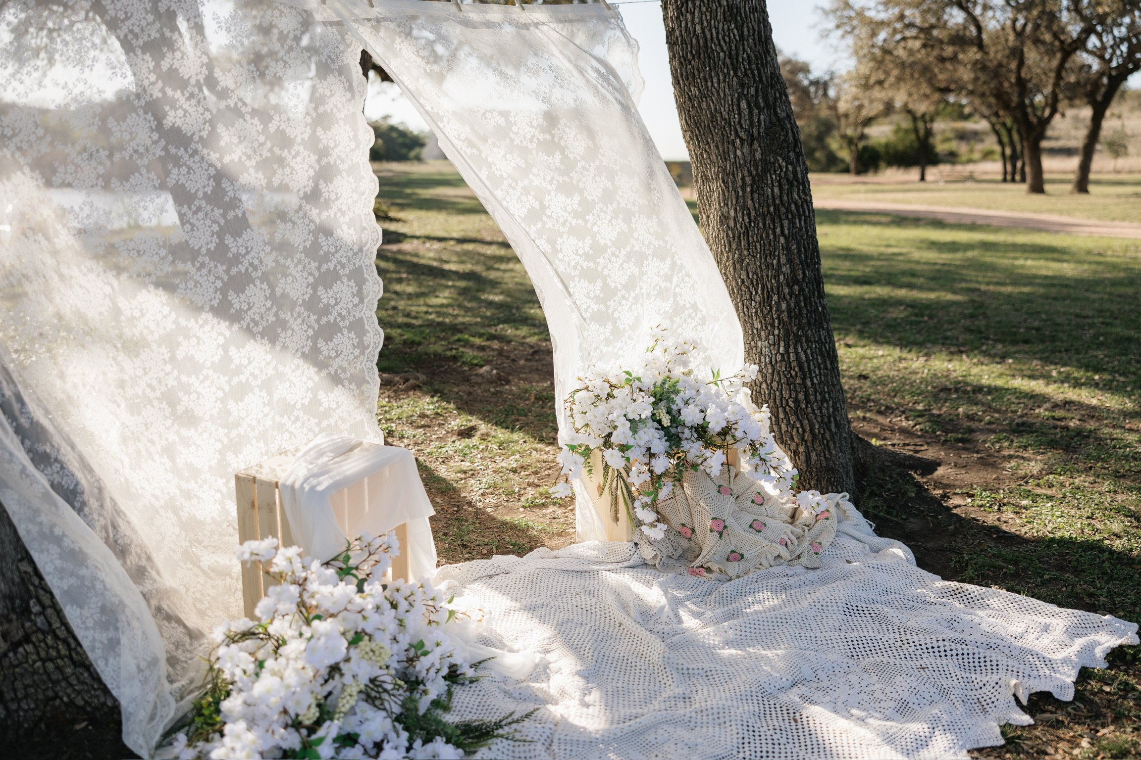 A wedding setup outdoors under trees with white lace draped fabric, floral arrangements, and a small wooden table, in a park-like setting.