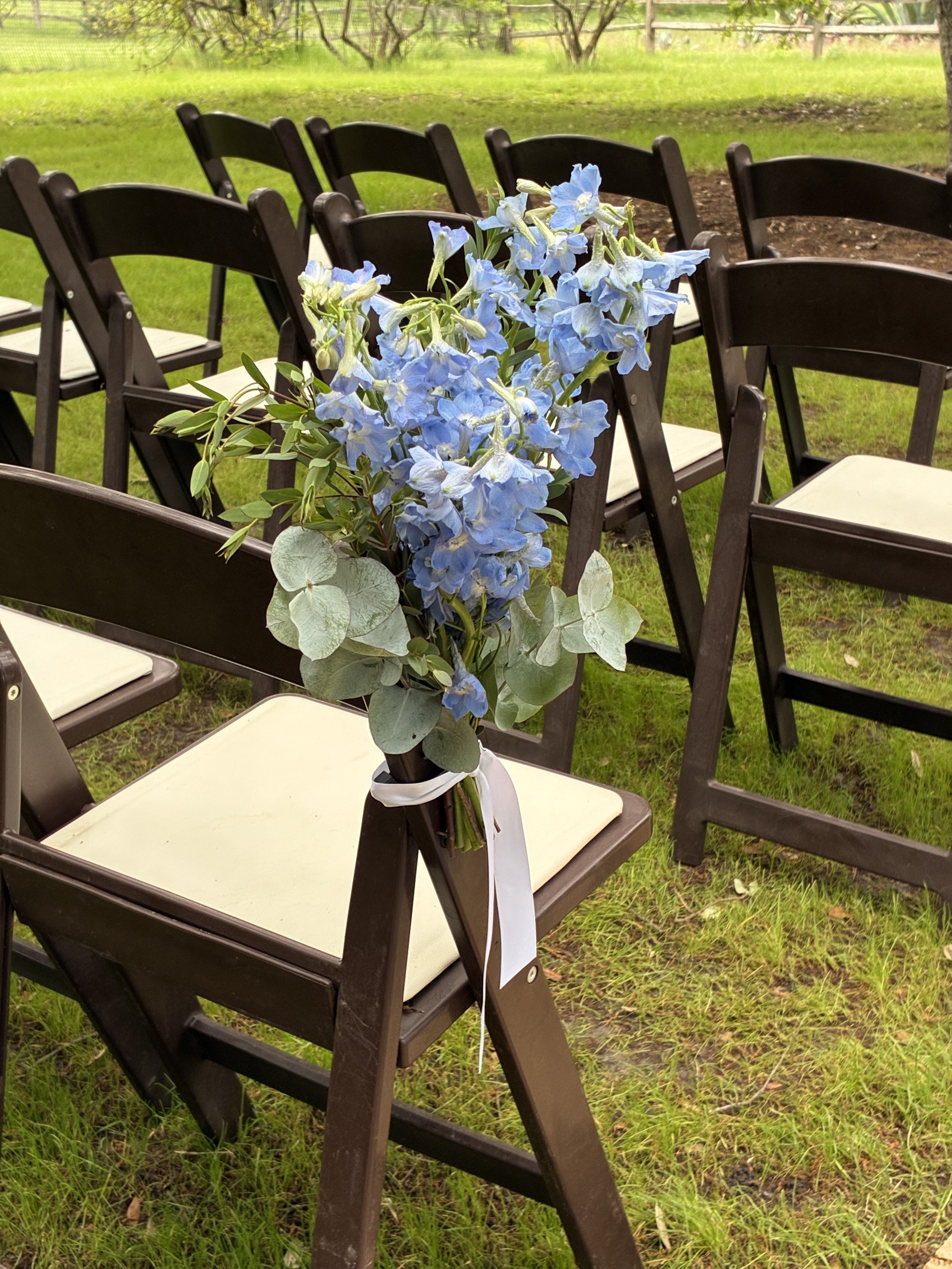 A bouquet of blue flowers with green leaves and accents of grayish leaves is attached to a black folding chair at an outdoor event, with more black chairs arranged in rows  for a wedding ceremony aisle on green grass visible in the background.