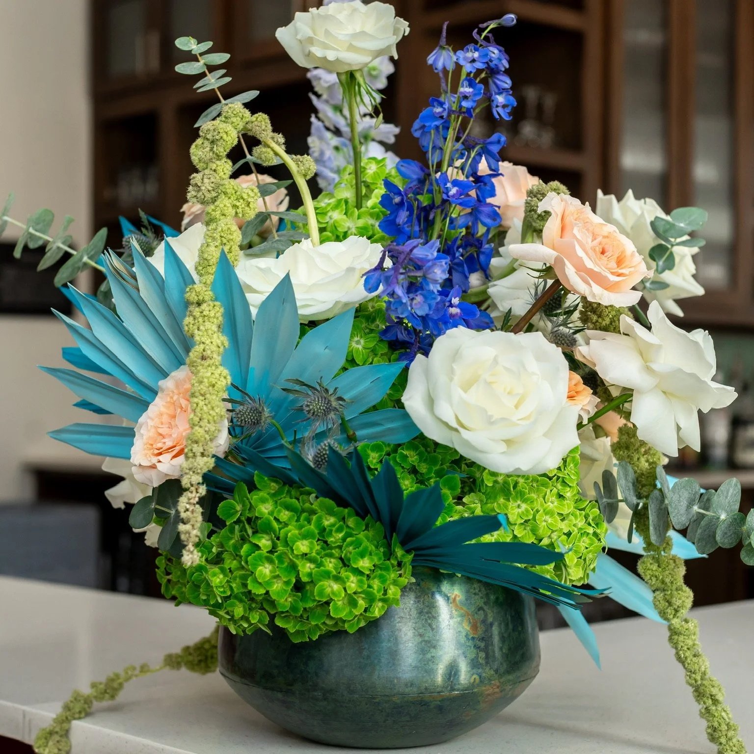 A floral arrangement in a dark ceramic vase containing white roses, peach roses, blue delphiniums, green hydrangeas, eucalyptus, and other greenery and flowers.