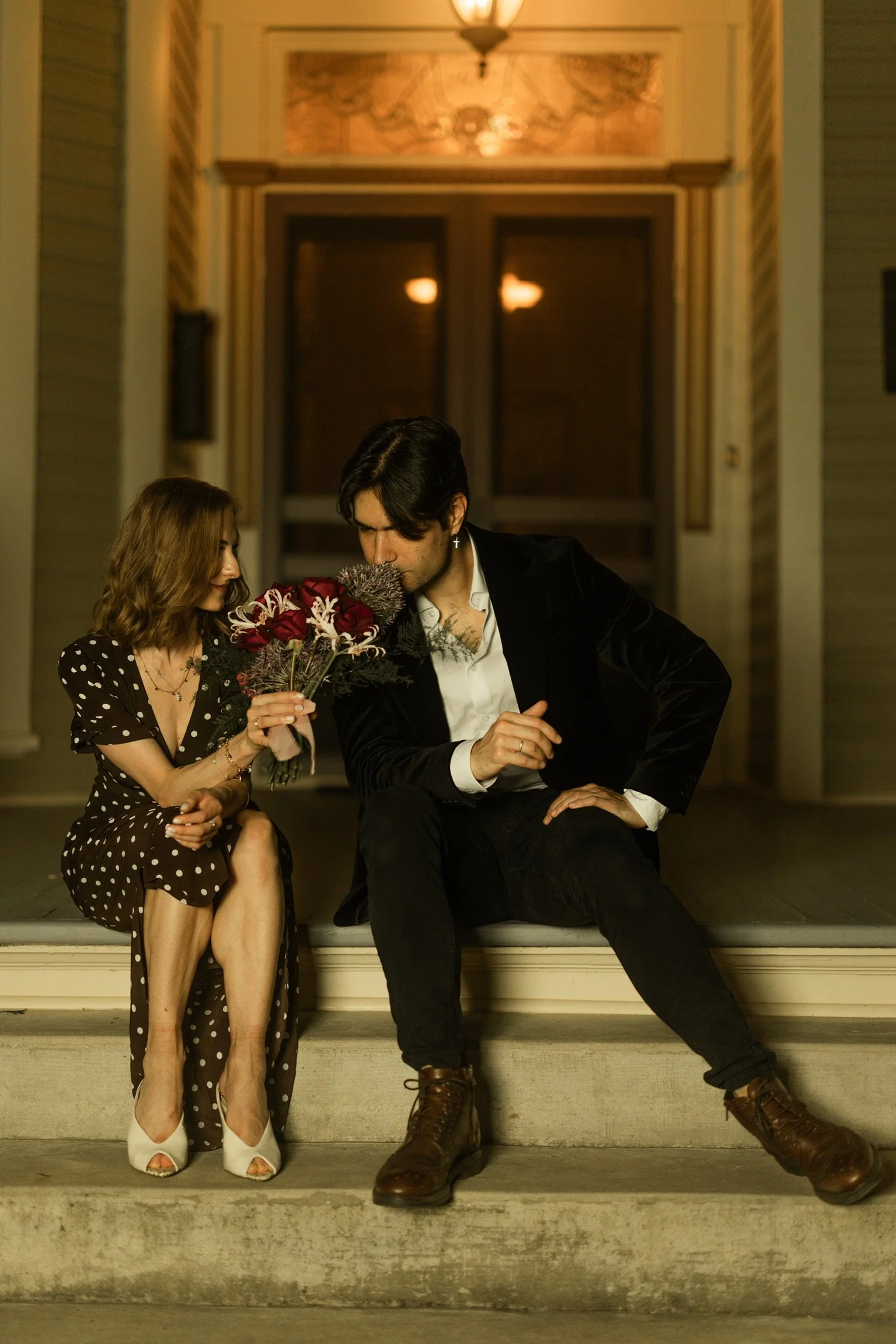 A man and woman sitting on front steps outside a house at night. The woman is holding a bouquet of flowers, wearing a polka dot dress and white heels, while the man, dressed in a black blazer, white shirt, and dark pants, looks at her.