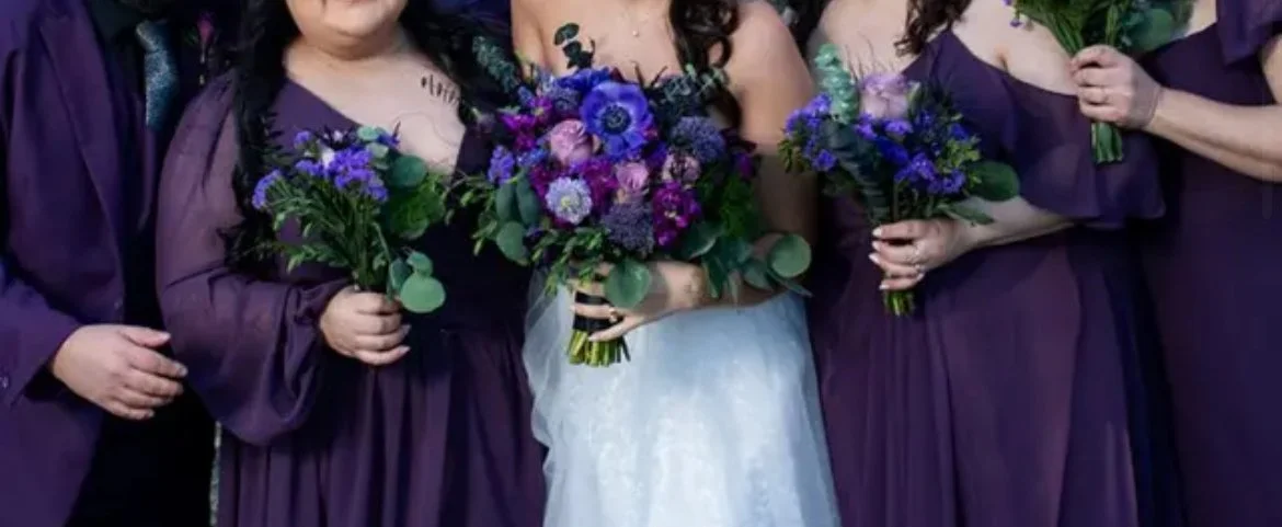 Close-up of a bride in a white dress holding a bouquet of purple and blue flowers, surrounded by bridesmaids in purple dresses holding matching bouquets.