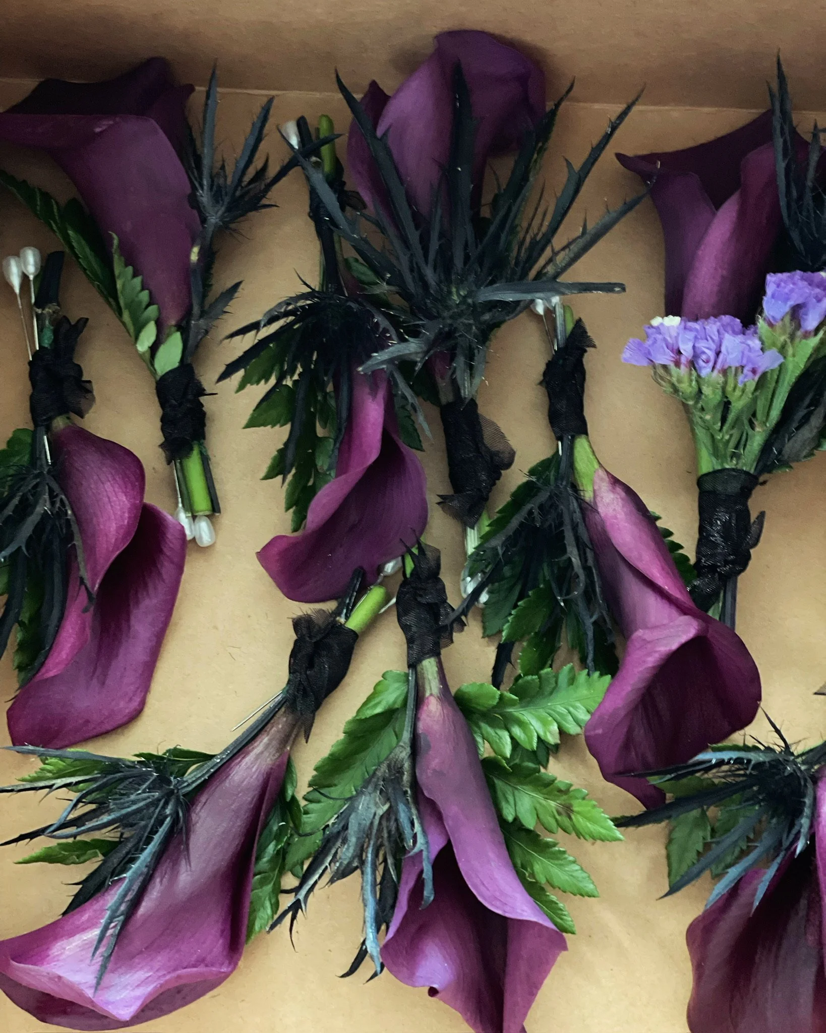 A collection of boutonnieres made with purple calla lilies, purple statice, green leaves, and black thistle, all tied with black ribbon.