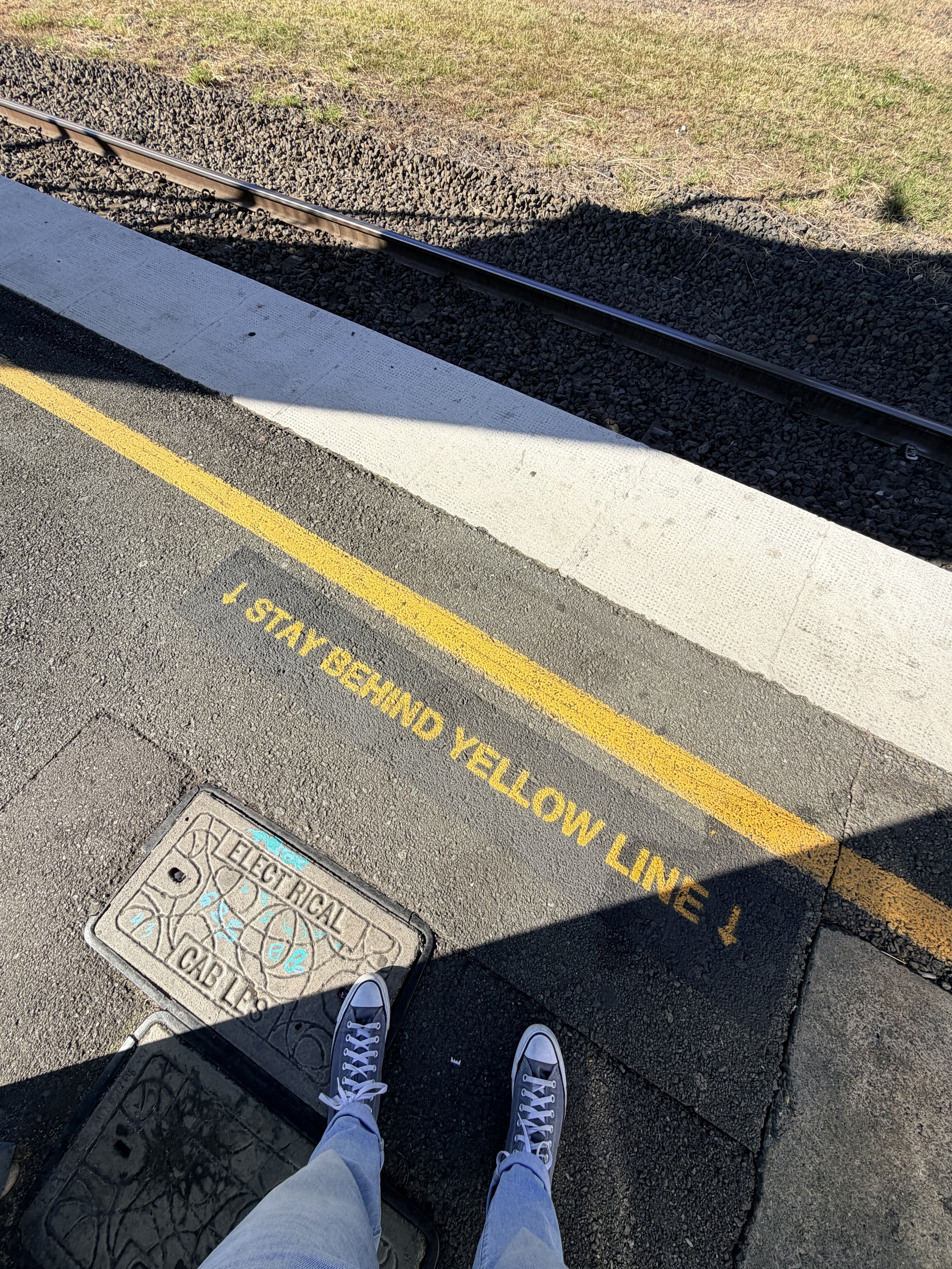 A person's feet wearing sneakers on a sidewalk near a yellow painted line and a manhole cover with blue chalk markings, with visible shadow and grass in the background.