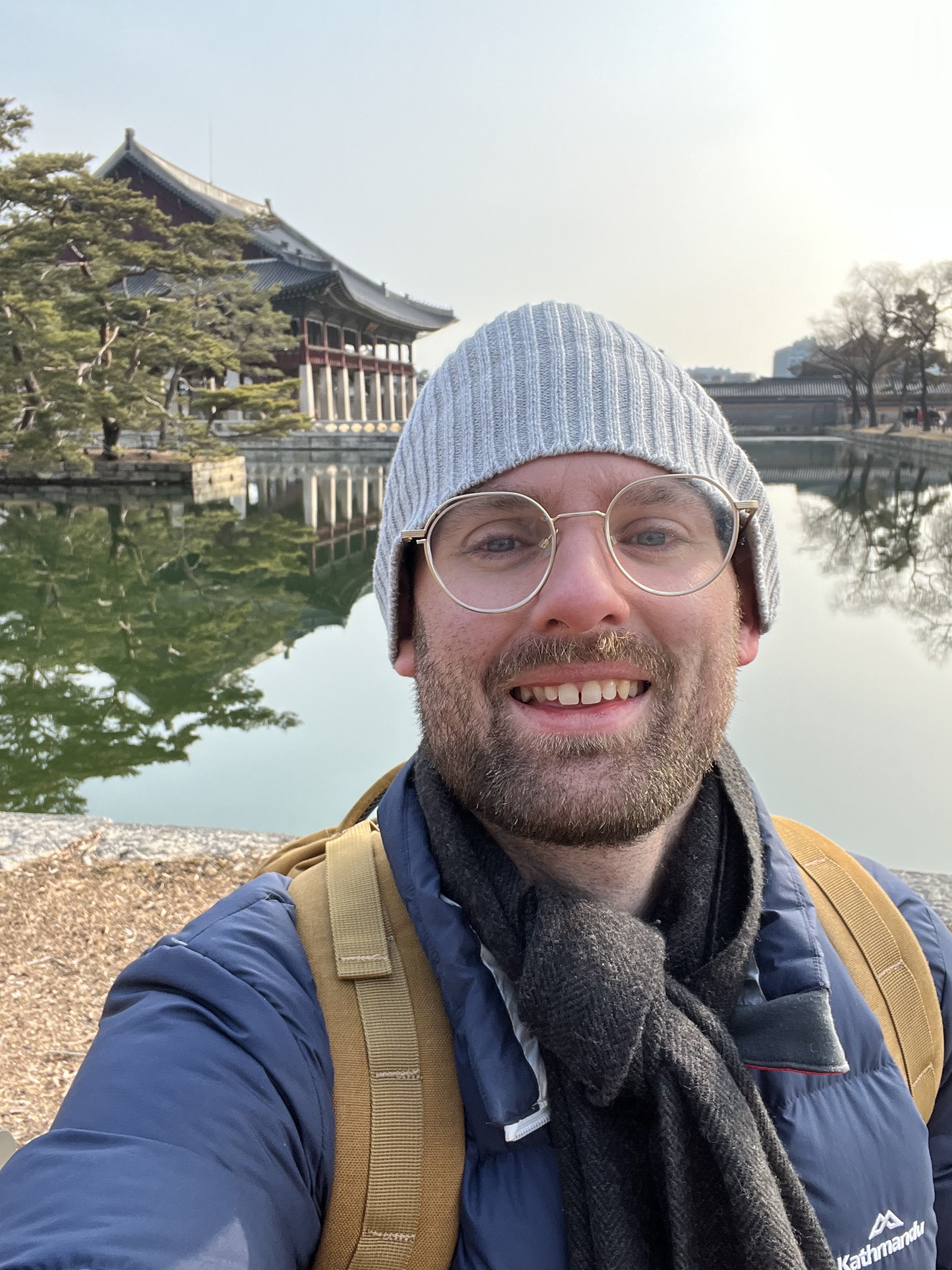 A smiling man taking a selfie outdoors near a traditional Asian-style building by a body of water, wearing a gray beanie, glasses, a blue jacket, and a backpack.