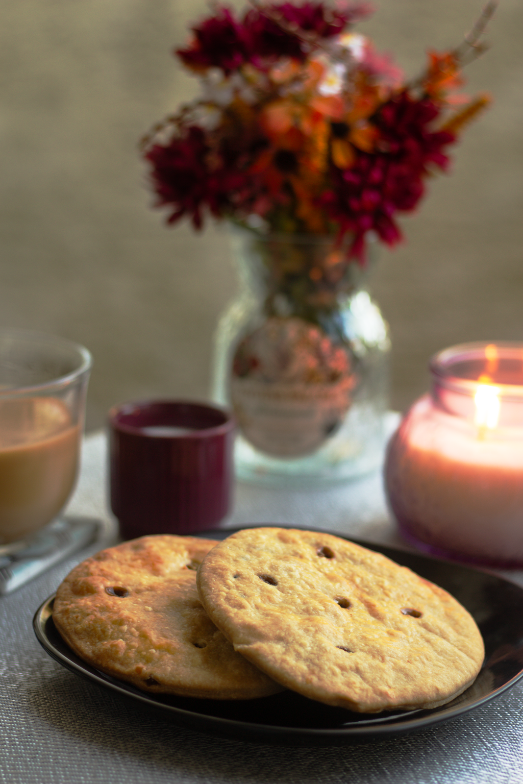 Plate with two cornbread muffins, a lit candle, a glass of coffee, and a small candle in a red holder, with a vase of red and purple flowers in the background on a table.