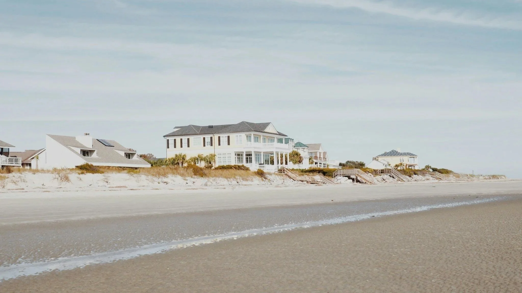 Row of beachfront houses along the shoreline with sandy beach and gentle waves in the foreground, under a cloudy sky.