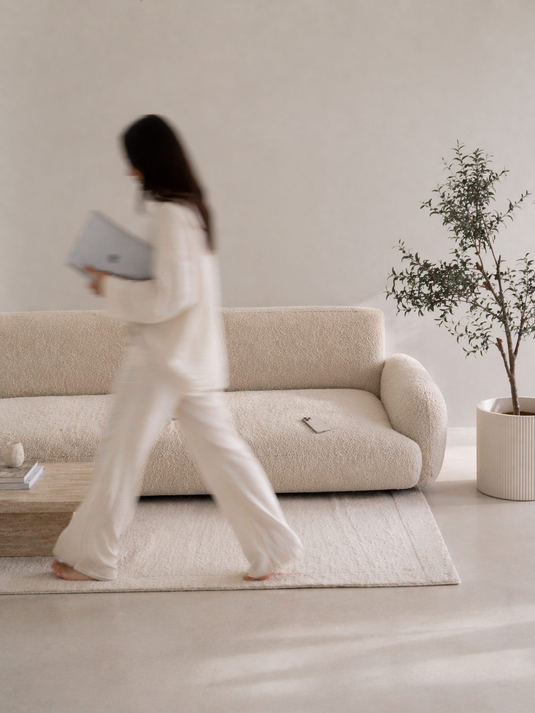 A woman in white pajamas walking past a beige couch in a minimalistic, neutral-colored room, carrying a laptop and with a cellphone on the couch. There is a potted plant in a white planter next to the couch.